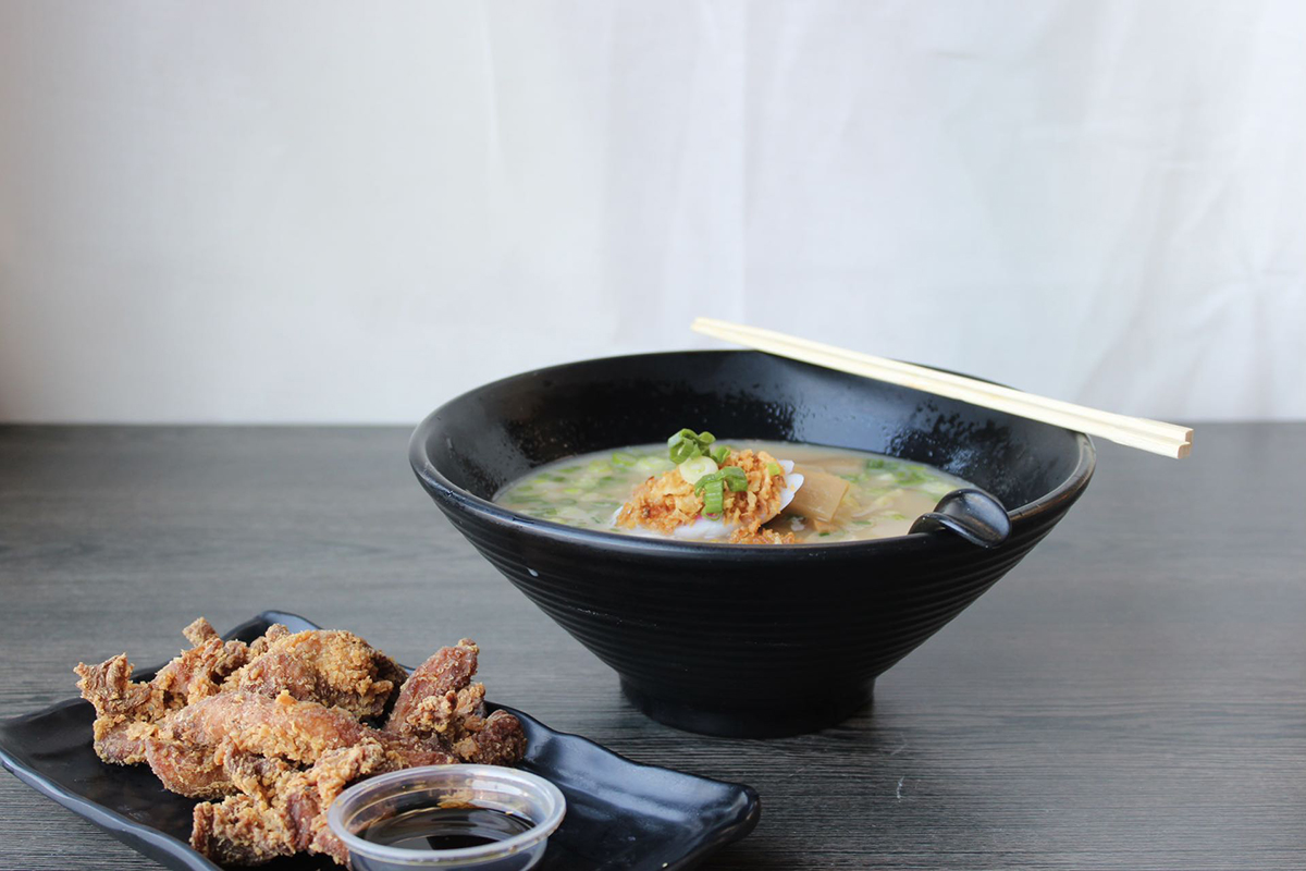 Ramen in black bowl topped with green onions and fried garnish, side plate of crispy fried chicken with soy dipping sauce, chopsticks resting on bowl rim, dark table against plain white background.