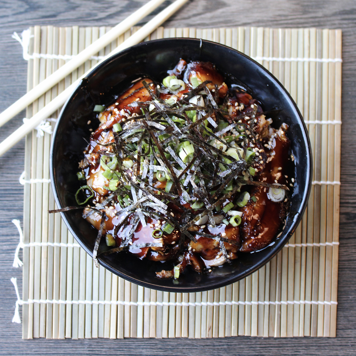 Top-down view of black bowl of unagi don, grilled eel glazed with teriyaki sauce over rice, topped with seaweed strips, green onions and sesame seeds, resting on bamboo mat with chopsticks.