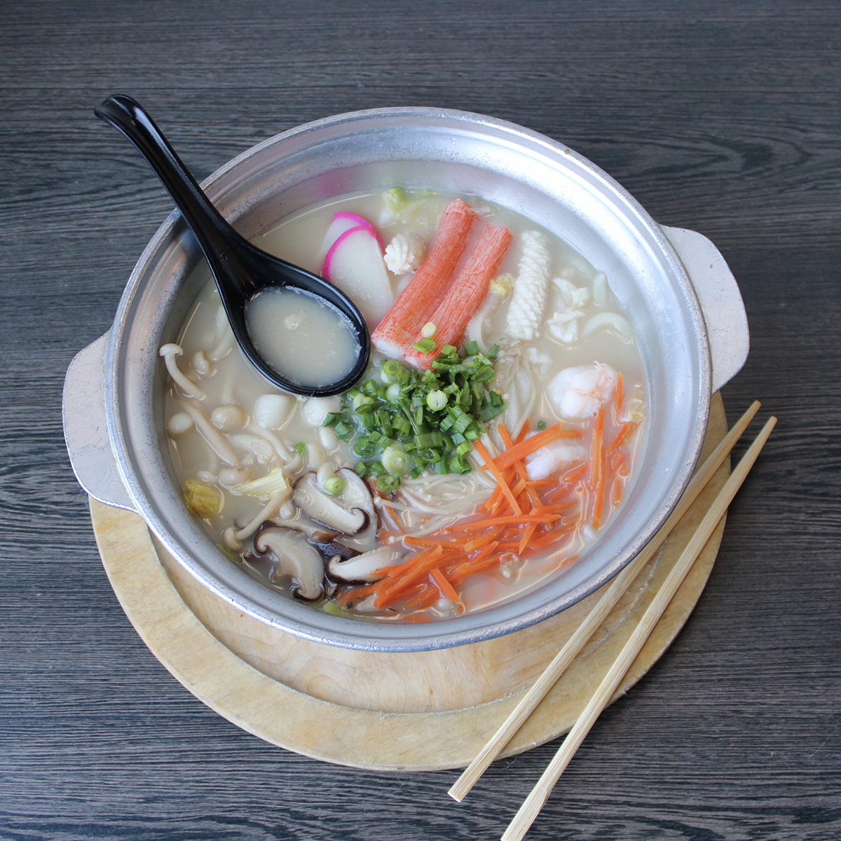 Top-down view of seafood hot pot in silver metal pot, creamy soup with shrimp, crab stick, mushrooms, carrot strips and green onions, black soup spoon and chopsticks on grey wooden table.