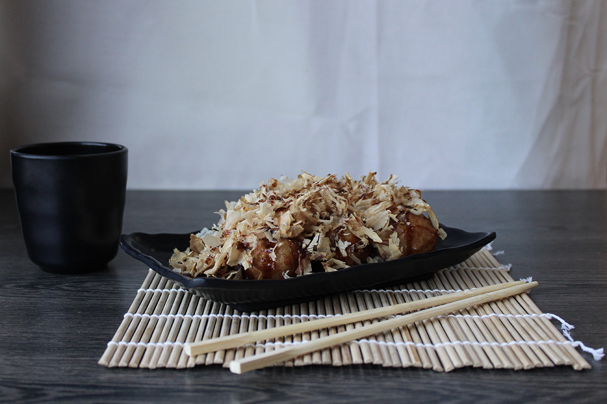 Takoyaki octopus balls piled on a black rectangular plate, generously covered with bonito flakes, served on bamboo mat with wooden chopsticks and black teacup on dark wooden table.