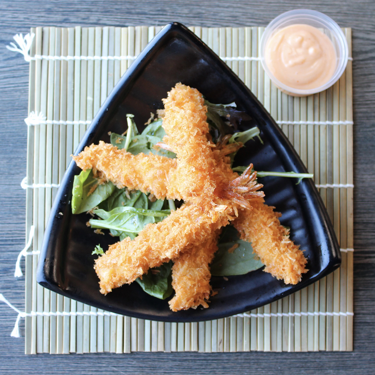 Top-down view of breaded tempura shrimp stacked on mixed salad greens in a black triangular plate, with small plastic cup of creamy orange sauce on bamboo placemat.