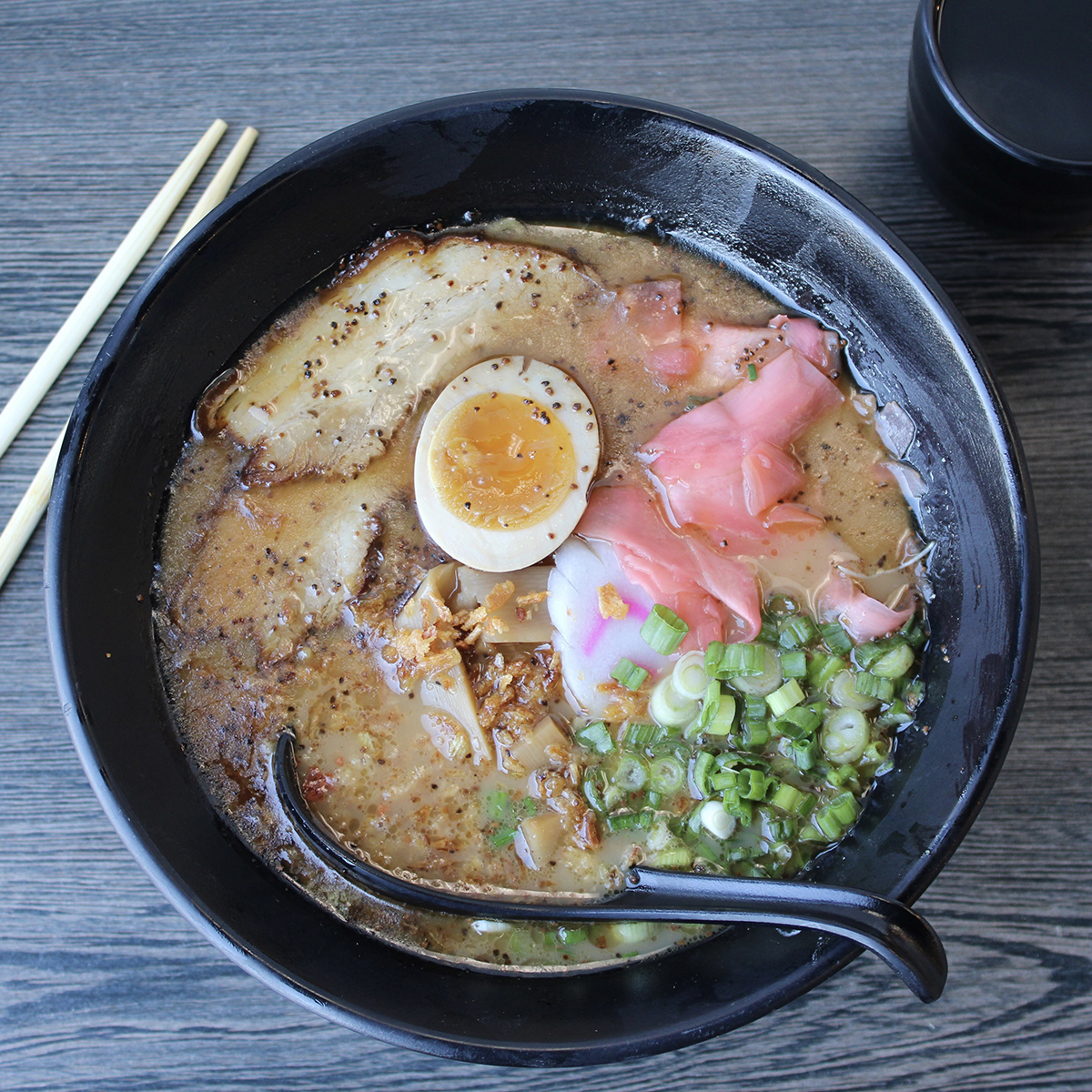 Top-down view of tonkotsu ramen in a black bowl, topped with chashu pork, runny soft boiled egg, pickled ginger, green onions, bamboo shoots, on grey wood table with chopsticks.