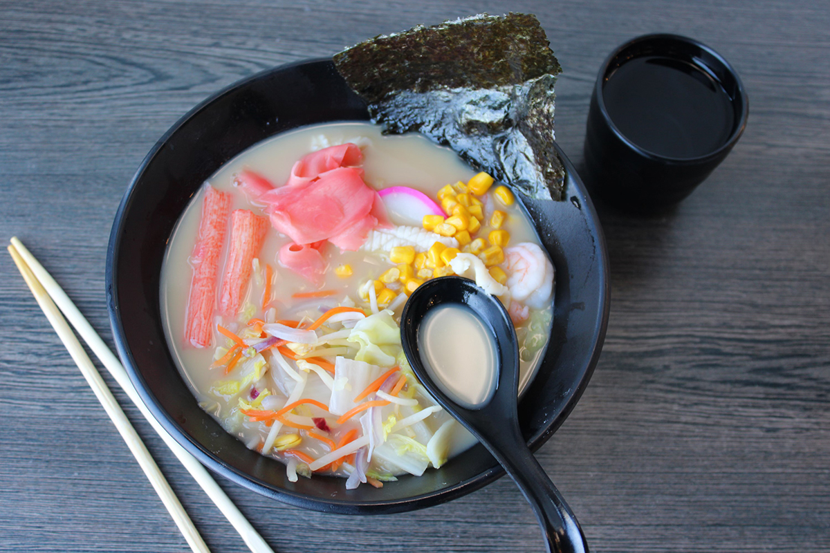 Top-down shot of seafood ramen in black bowl with creamy broth, shrimp, crab stick, corn, pickled ginger, seaweed and mixed vegetables, chopsticks and black cup on grey wooden table.