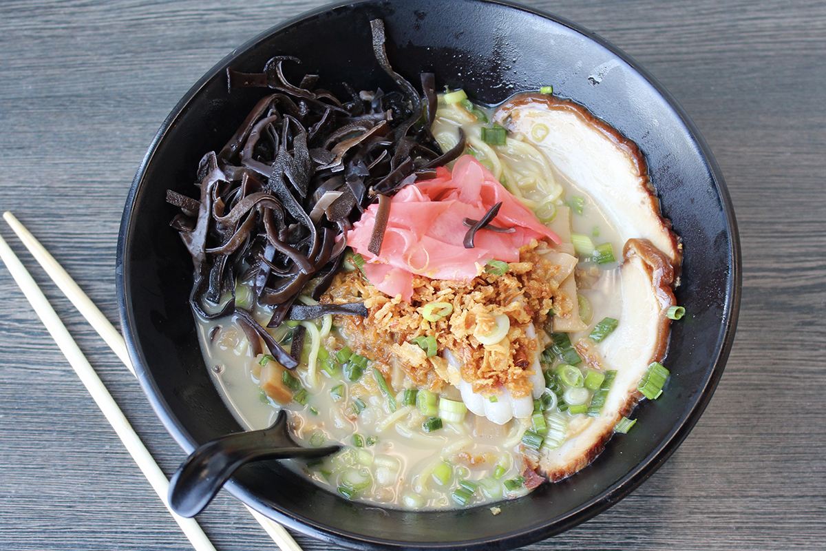 Top-down view of creamy tonkotsu ramen in a black bowl, topped with chashu pork, wood ear mushrooms, pickled ginger, fried onion and green onions, placed on grey wood table with chopsticks.