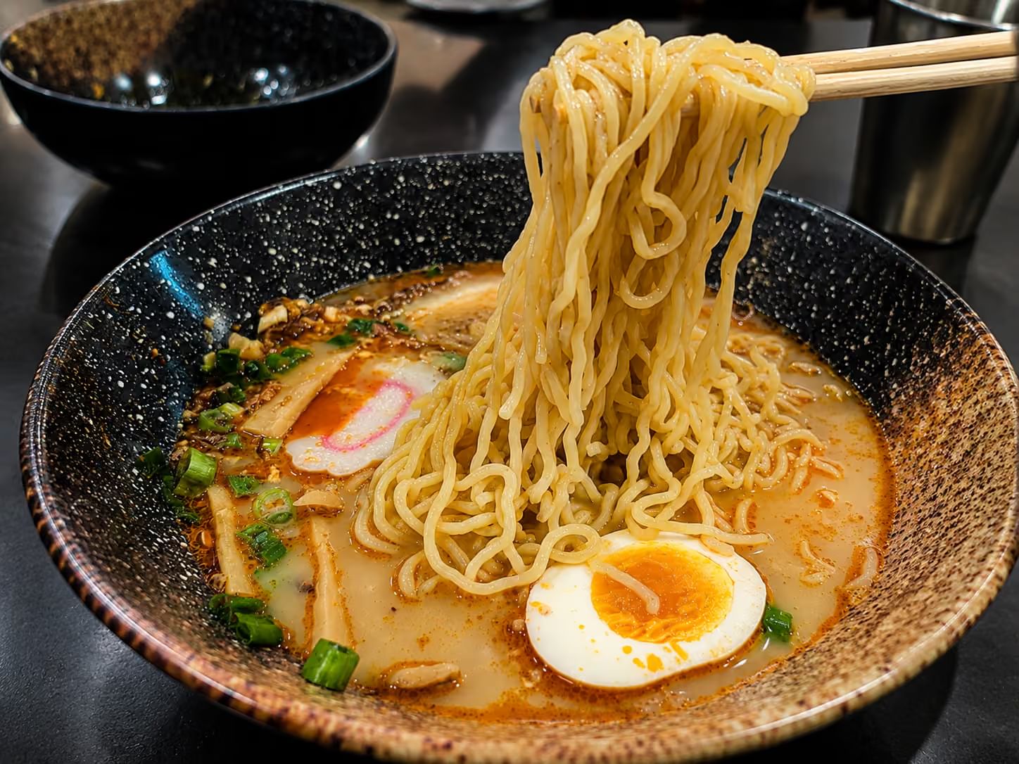 Tonkotsu ramen in speckled ceramic bowl, chopsticks lifting noodles, topped with soft boiled egg, narutomaki, bamboo shoots and green onion