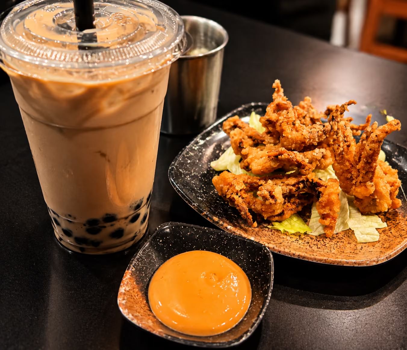 Plastic cup of bubble tea on black table, plate of crispy fried squid tentacles on lettuce, small dish of orange dipping sauce