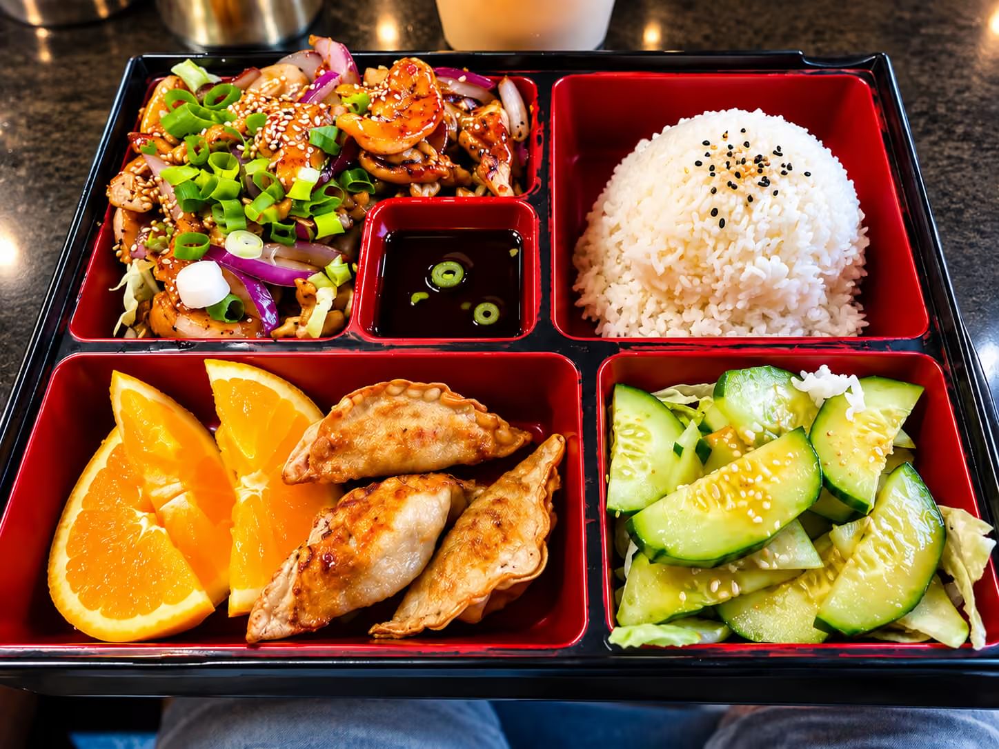 Red divided bento tray with steamed rice, pan-fried gyoza, shrimp chicken stir fry, cucumber salad, orange slices and soy sauce
