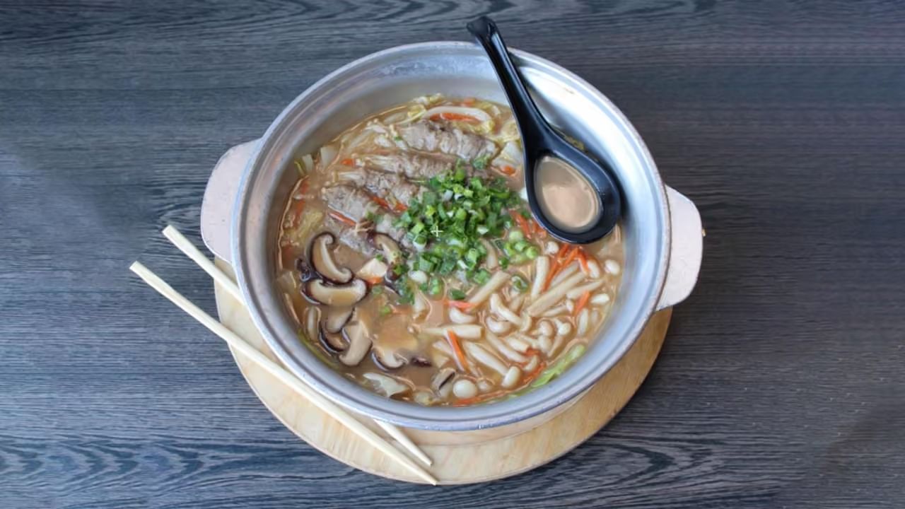 Top-down view of beef udon hot pot in aluminum pot, shiitake, carrot, cabbage and green onion, wooden chopsticks and black soup spoon on wood table