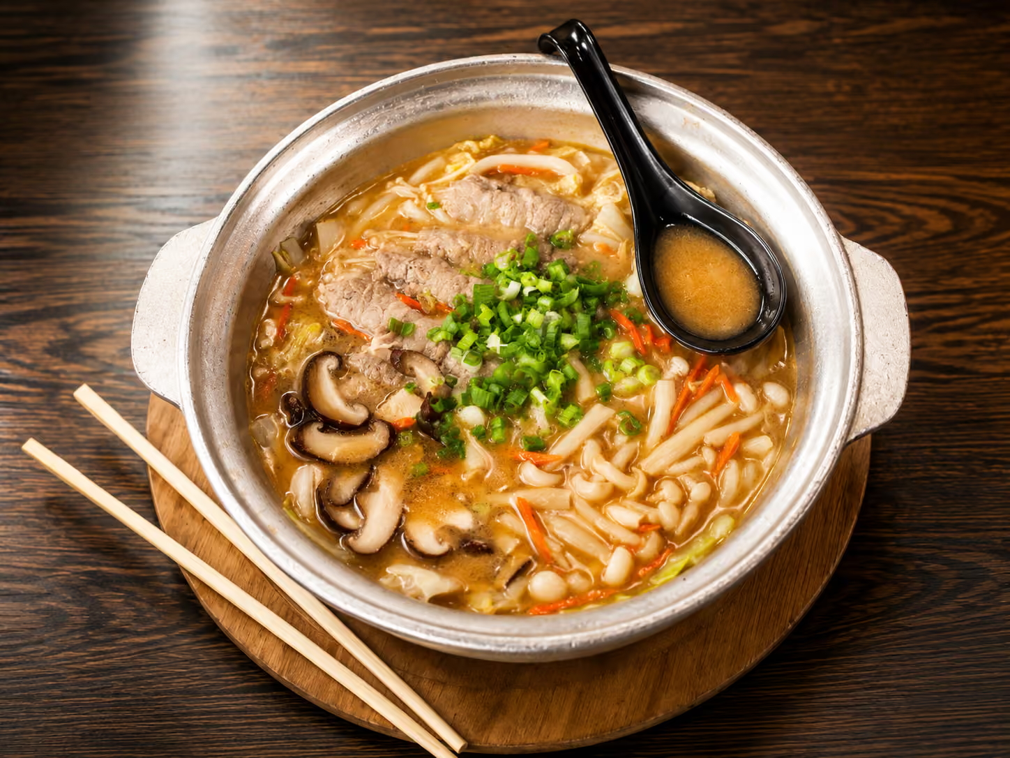 Aluminum pot of beef udon hot pot with shiitake, carrot, cabbage and green onion, wooden chopsticks and black soup spoon on dark wood table