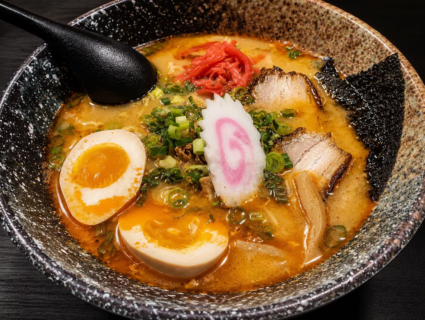 Rich tonkotsu ramen in speckled ceramic bowl, topped with soft-boiled eggs, chashu pork, narutomaki, green onion, ginger and nori, with black soup spoon
