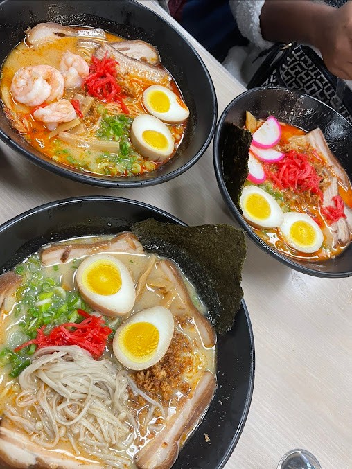 Three black bowls of Japanese ramen, creamy tonkotsu and spicy broth, topped with chashu pork, shrimp, soft boiled eggs, red ginger, green onion and nori seaweed on a light table.