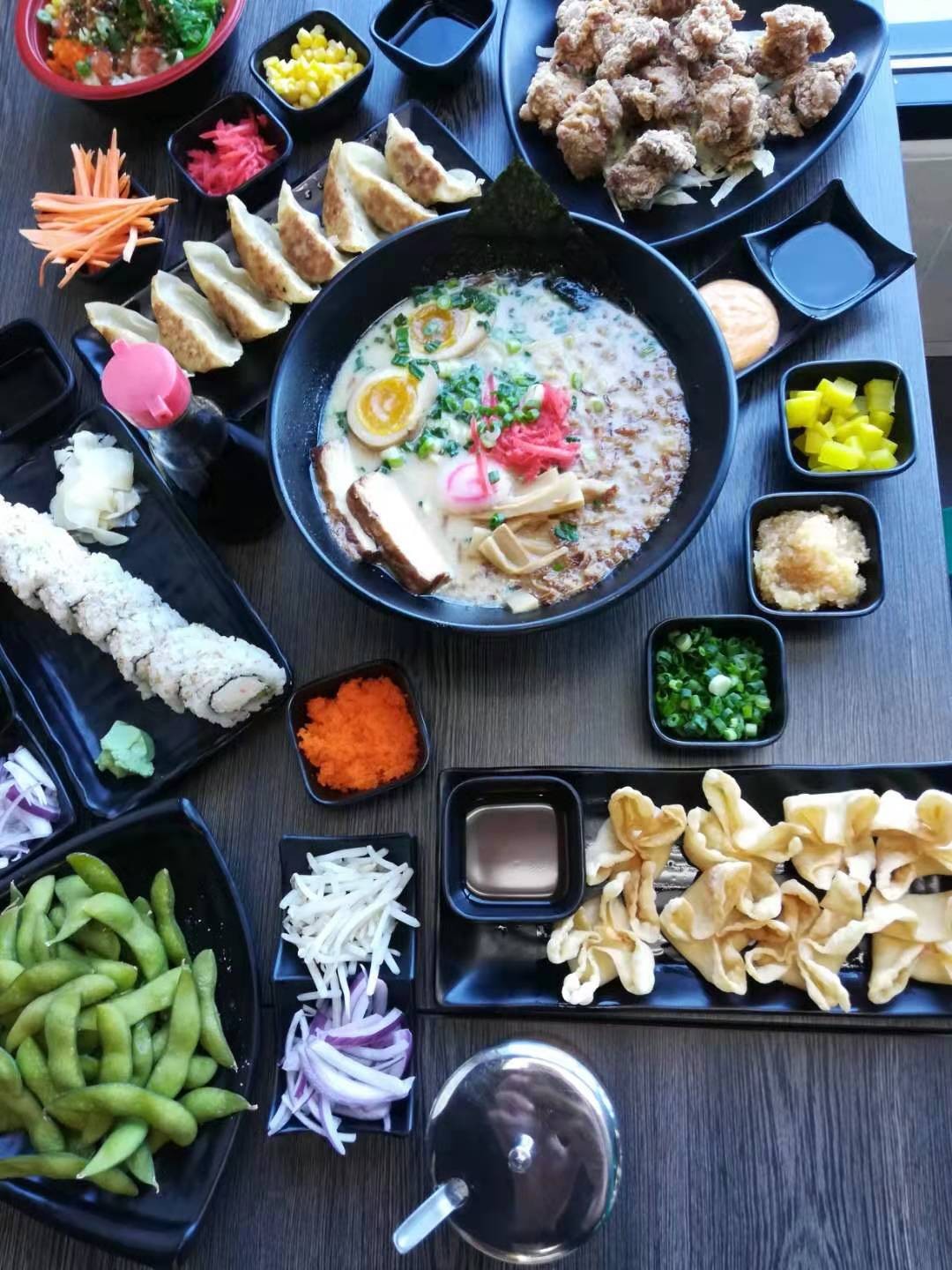 Table spread of assorted Japanese dishes: tonkotsu ramen, gyoza, sushi roll, edamame, fried chicken, tempura and multiple small side dishes on dark plates.