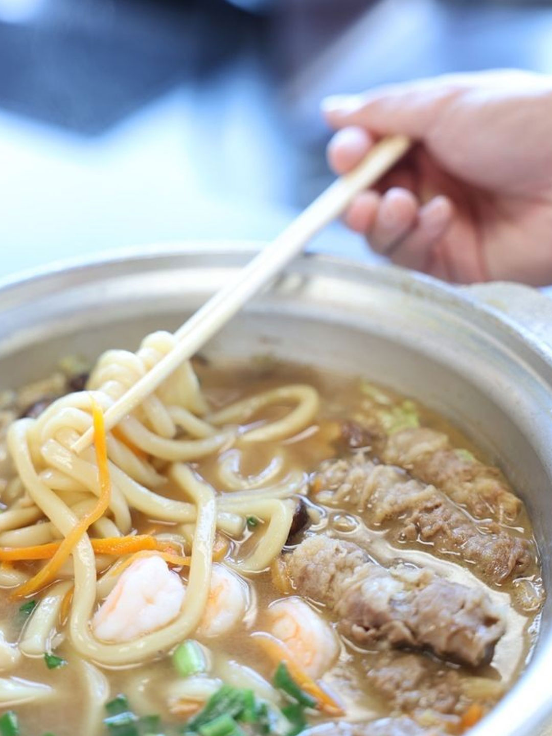 Metal hot pot with hearty udon noodle soup, topped with sliced beef, shrimp, green onions and shredded carrot; chopsticks lifting thick udon noodles.