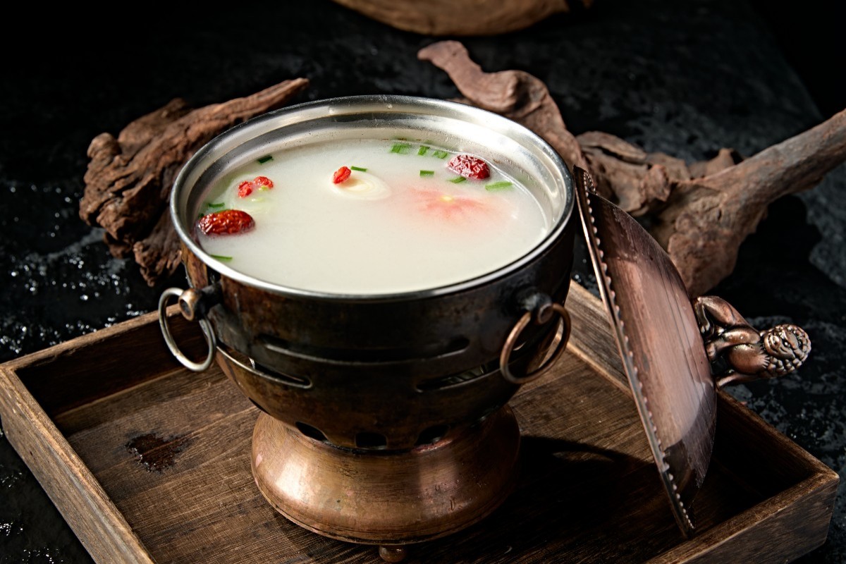 A light-colored soup in a rustic metal pot with red dates, green scallions, and other garnishes, placed on a wooden tray against a dark background.