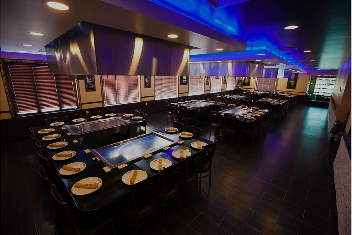 Spacious dimly lit dining room with lined teppanyaki tables pre-set with plates and blue ceiling lighting at Akira, a Japanese Restaurant in Glenmont