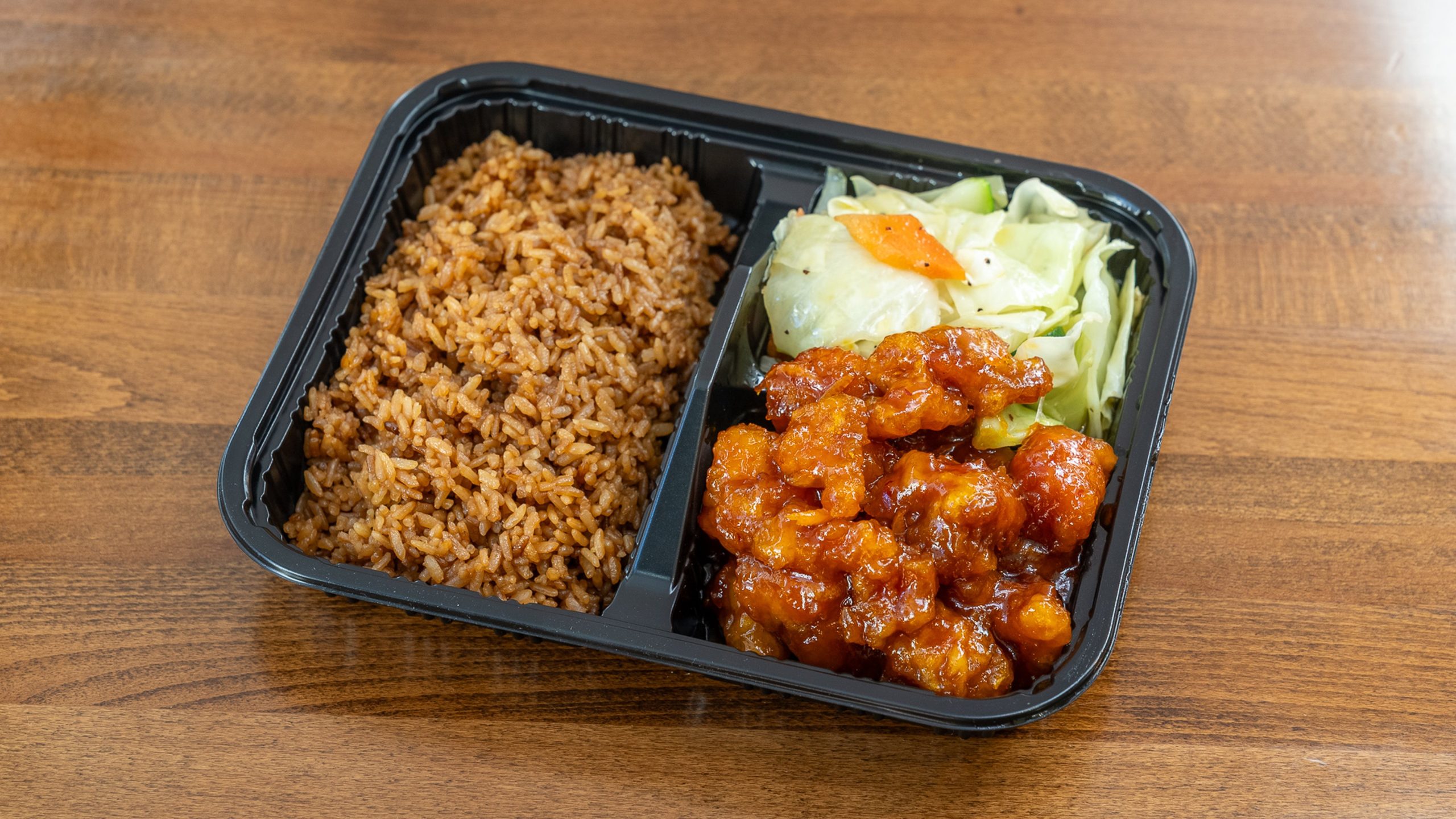 Black plastic two‑compartment takeout tray on wooden table. Left side filled with brown fried rice; right side holds stir‑fried cabbage with carrot slice above glossy orange‑sauced crispy chicken chunks.