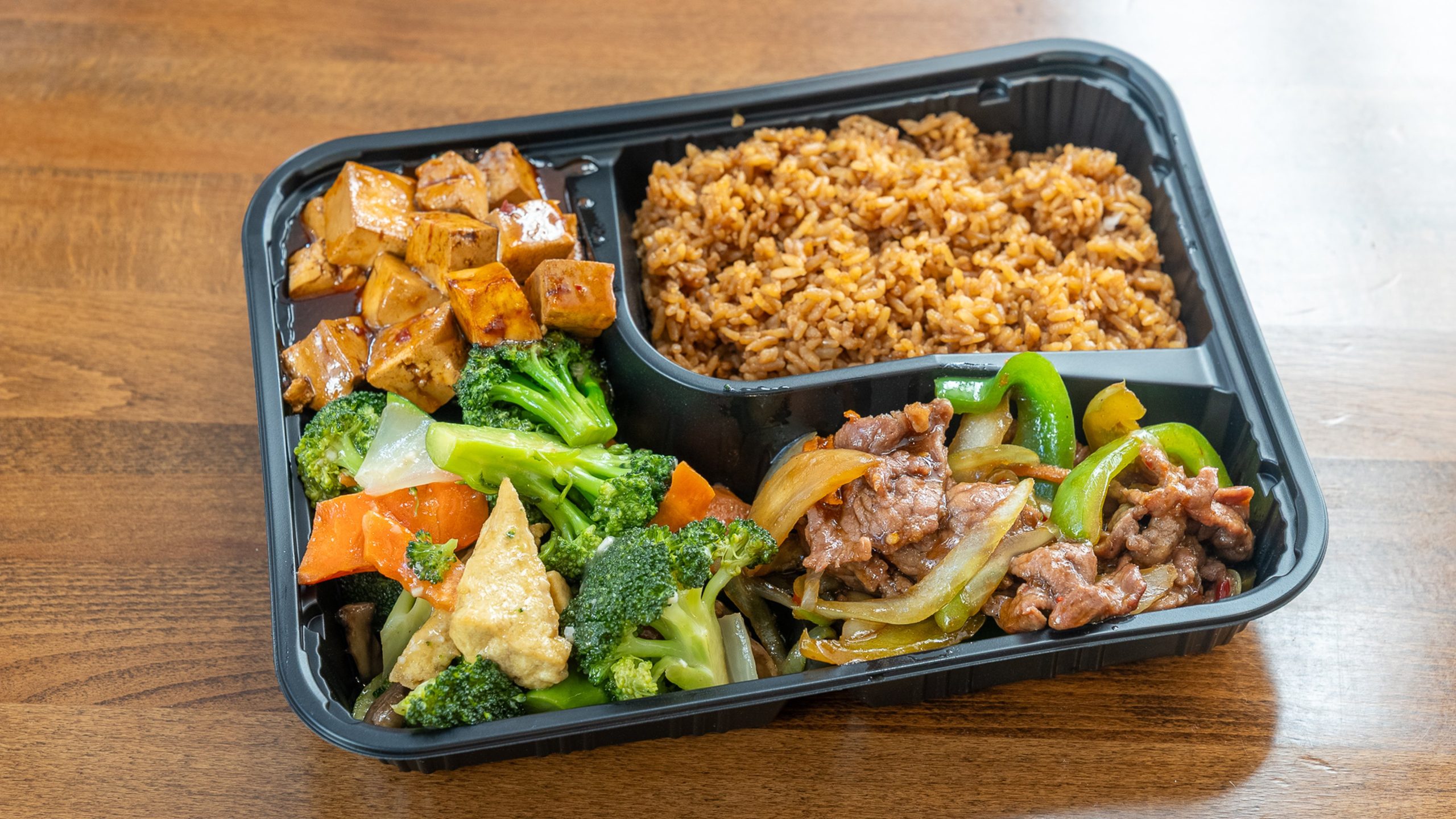 Black three‑compartment takeout container. Top‑left cubed braised tofu in dark sauce; top‑right brown fried rice; bottom section combines broccoli‑tofu stir‑fry and sliced beef sautéed with green peppers, onions and carrots.