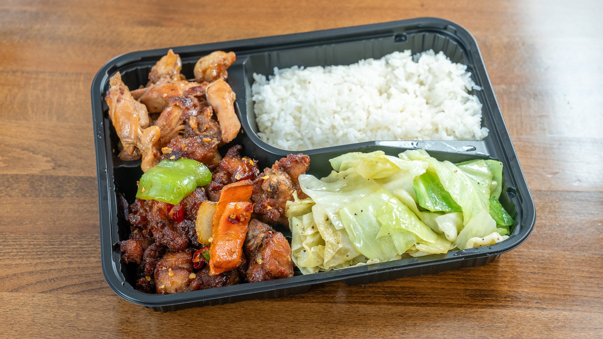 Black three‑section takeout tray on wood table. Top‑left light roasted chicken pieces; top‑right steamed white rice; bottom holds spicy dried‑chili chicken with green pepper and carrot alongside sautéed cabbage.