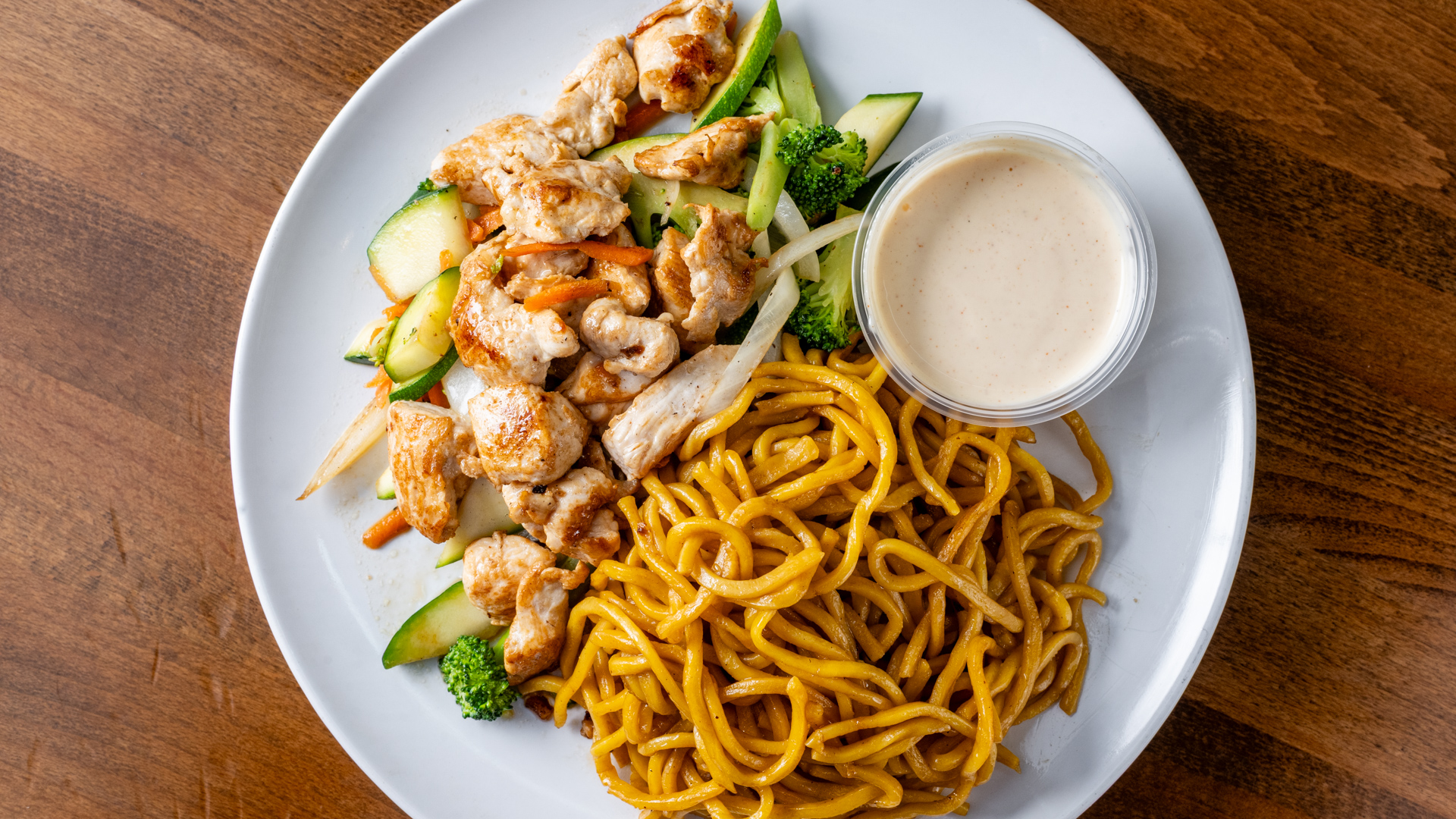 Overhead shot of white round plate. Grilled chicken breast chunks with zucchini, broccoli, carrot and onion on upper half; golden stir‑fried lo mein noodles on lower half; small clear cup of creamy white yum‑yum sauce on the side.