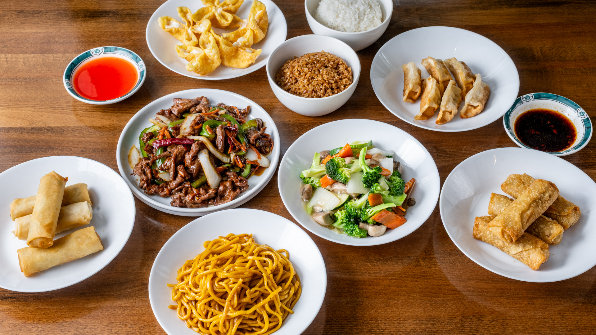 Wooden table full‑spread combo meal. Center plates: pepper‑beef stir‑fry with green peppers and onions, mixed broccoli‑mushroom vegetables. Surrounding bowls: white rice, fried rice, lo mein; plates of crab rangoon, pan‑fried dumplings, egg rolls with sweet‑and‑sour and chili dipping sauces.
