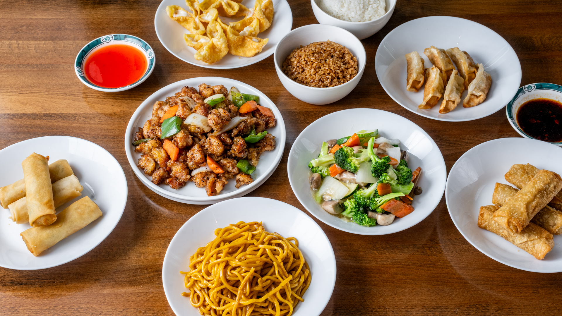 Overhead wooden‑table combo spread. Center: black‑pepper chicken stir‑fry with green peppers, onions, carrots and visible pepper flakes, plus broccoli‑mushroom vegetable medley. Surrounding: white rice, fried rice, lo mein, crab rangoon, pan‑fried dumplings, egg rolls with red sweet‑sour and dark chili sauces.