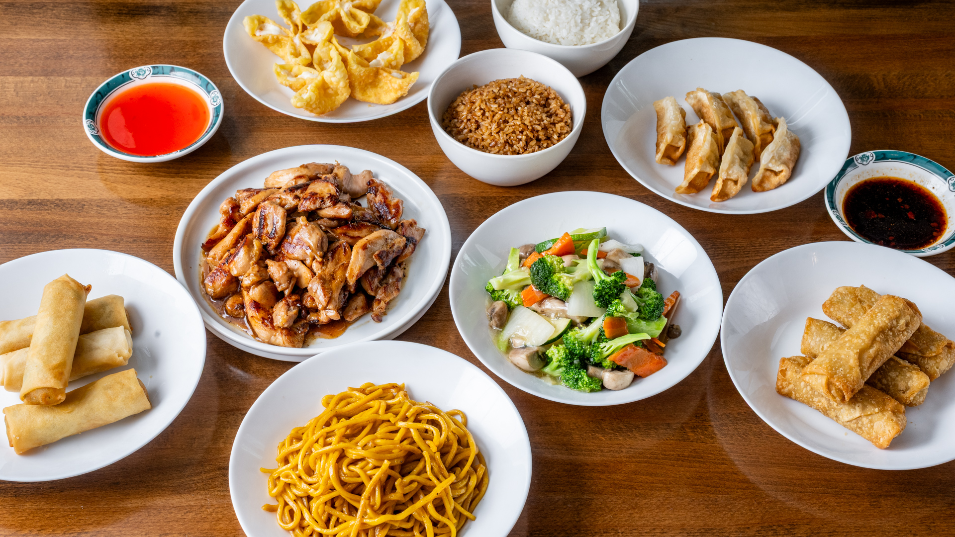 Wooden table full combo meal. Center: glazed teriyaki chicken pieces in shiny brown sauce alongside broccoli‑mushroom vegetable stir‑fry. Surrounding dishes: steamed white rice, fried rice, lo mein noodles, crab rangoon, pan‑fried dumplings, crispy egg rolls with sweet‑and‑sour and chili dipping sauces.