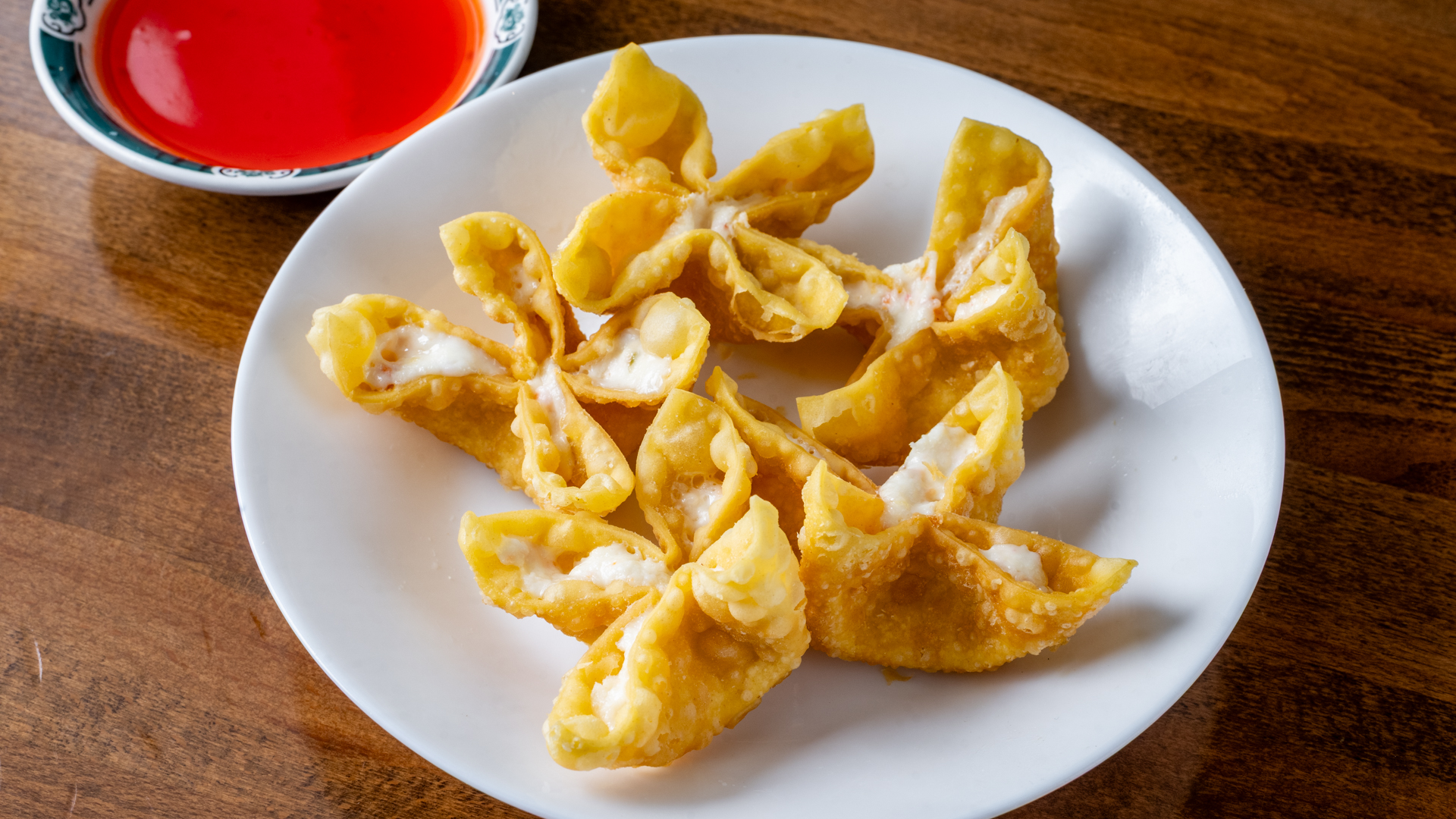 Close‑up of six golden fried crab rangoon wontons arranged on white round plate. Flower‑petal shaped crispy wrappers reveal creamy white crab‑cream‑cheese filling. Small patterned dish of bright red sweet‑and‑sour sauce in upper‑left on wooden table.