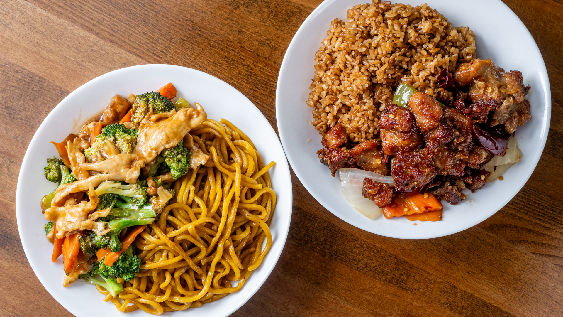 Two white oval plates side by side on wooden table. Left plate: chicken lo mein with broccoli and carrot in brown sauce. Right plate: brown fried rice topped with crispy spicy chicken pieces, dried red chilies, green pepper, onion and carrot slices.