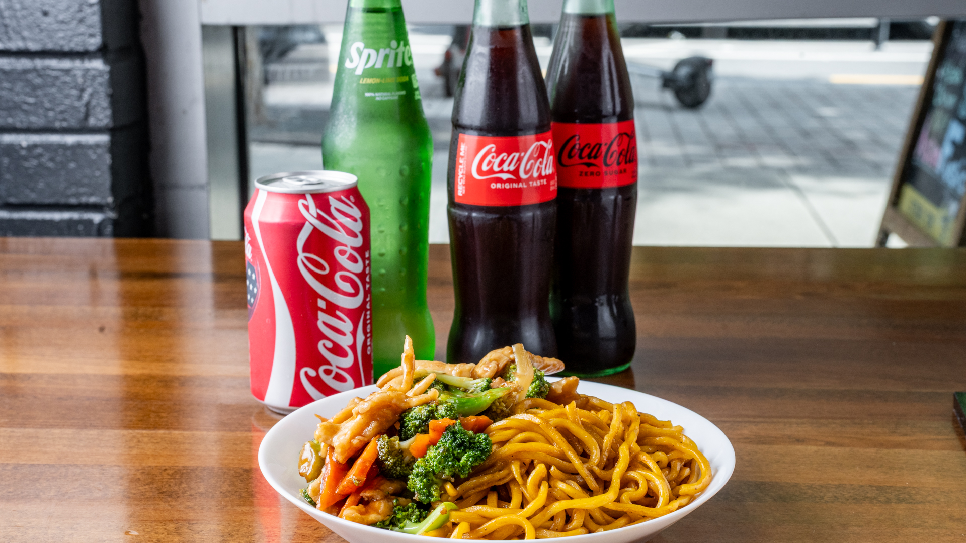Chicken broccoli lo mein on white plate in foreground. Behind it on wooden restaurant table: red Coca‑Cola can, green Sprite glass bottle, Coca‑Cola Original glass bottle and Coca‑Cola Zero Sugar glass bottle. Blurred storefront window and street in background.