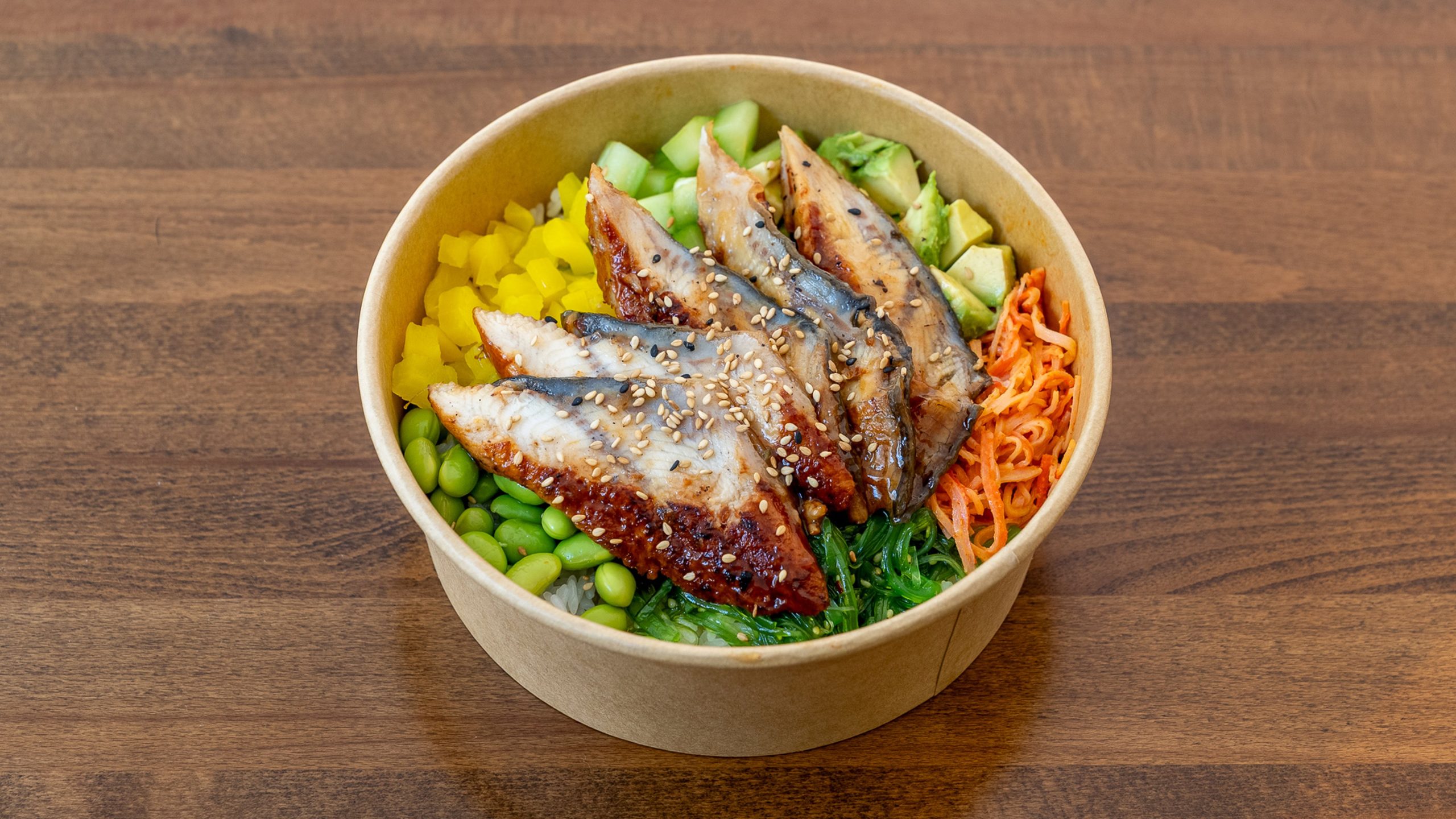 Top‑down view of unagi poke bowl in kraft paper bowl, glazed grilled eel slices sprinkled with sesame, mixed with edamame, avocado, cucumber, seaweed salad and diced bell pepper over rice on wooden table.