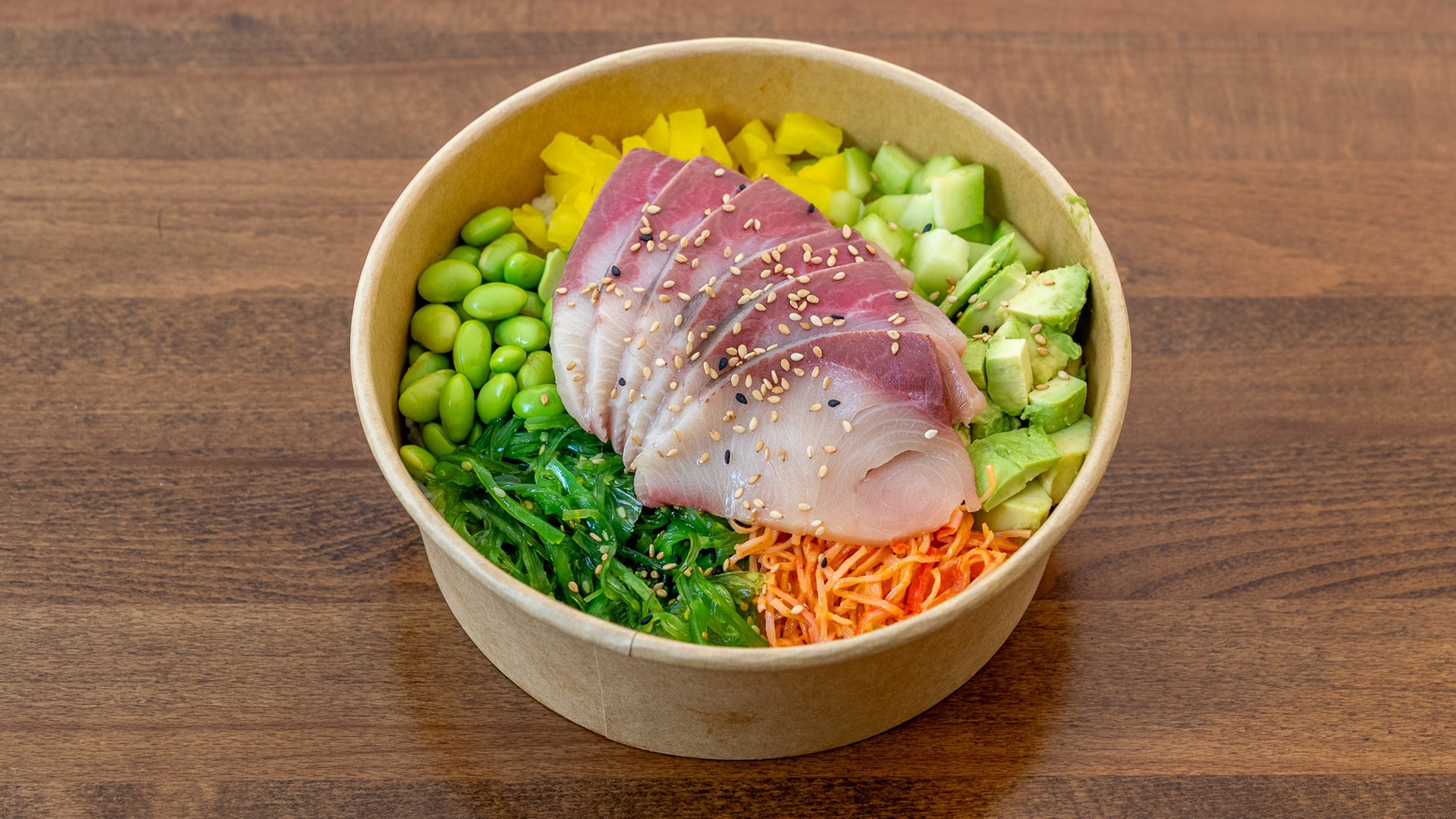 Top‑down shot of hamachi poke bowl in kraft paper bowl, raw yellowtail fish slices topped with sesame seeds, served with edamame, avocado, seaweed salad, carrot shreds and diced veggies on rice.