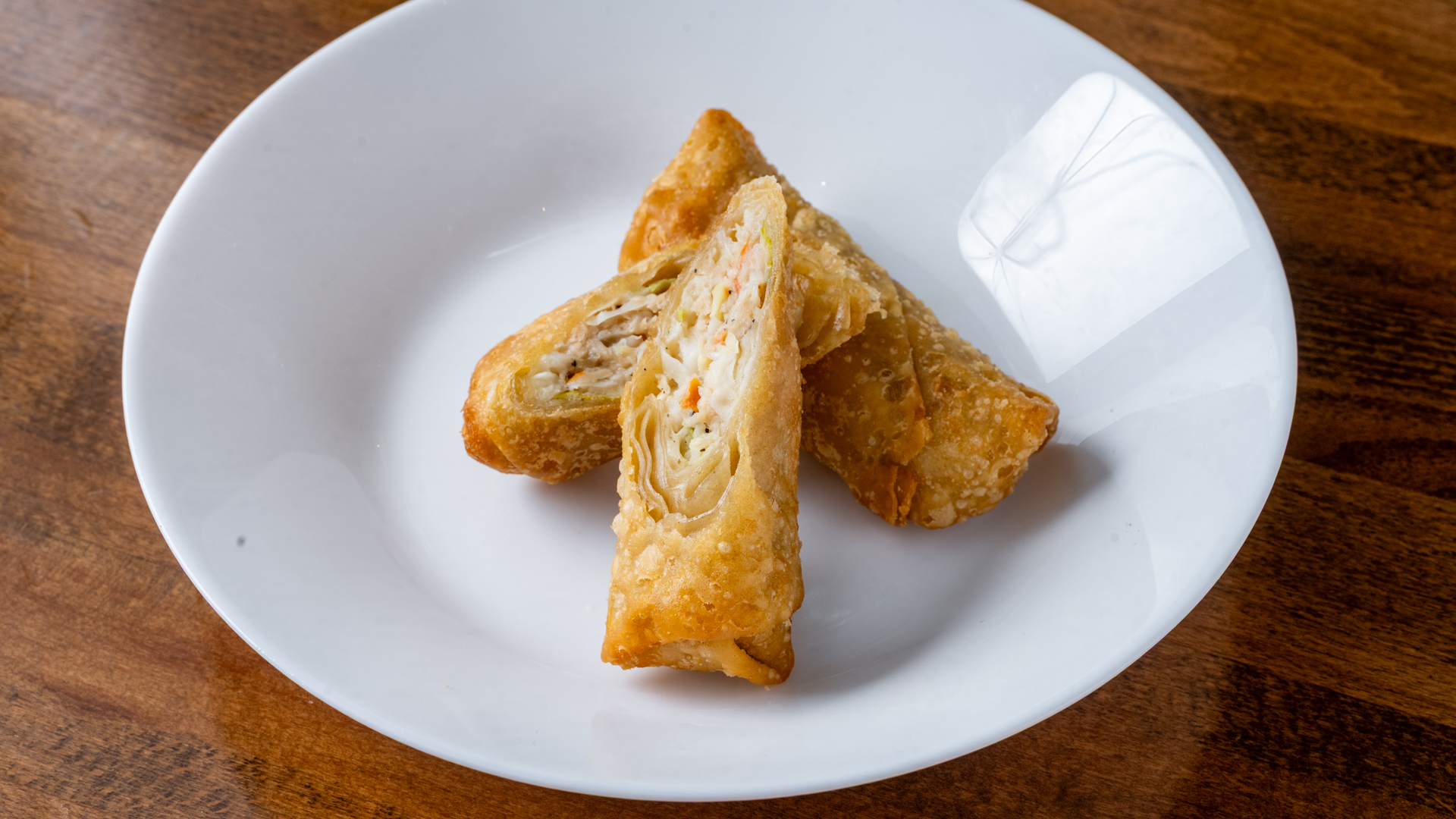 Three golden‑brown deep‑fried spring rolls on white ceramic plate, one cut open showing shredded cabbage, carrot and meat filling, placed on dark wooden table surface.
