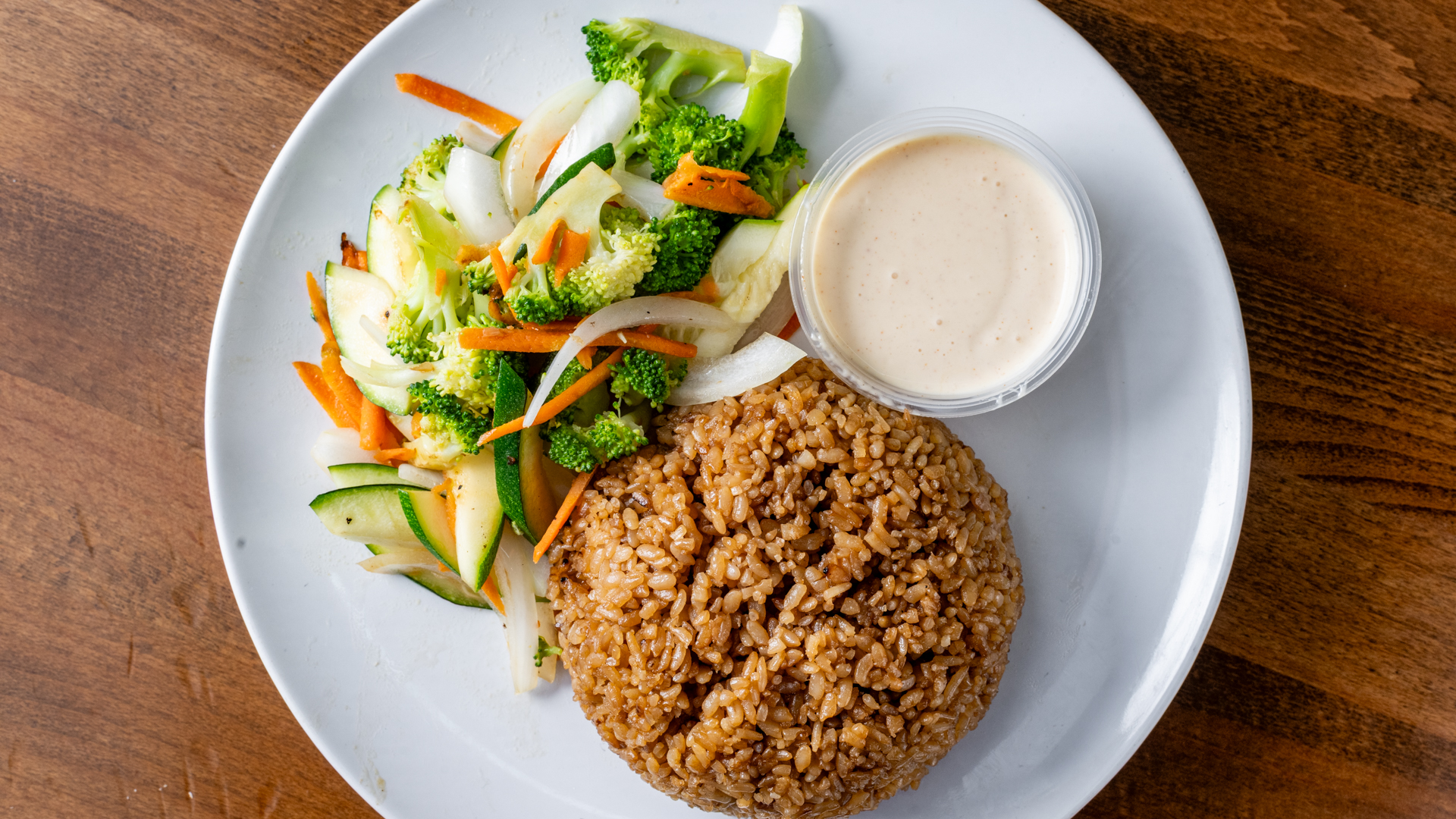 Overhead view of hibachi‑style fried rice mound on white plate, paired with stir‑fried broccoli, zucchini, onion, carrot and small container of creamy dipping sauce on wood table.