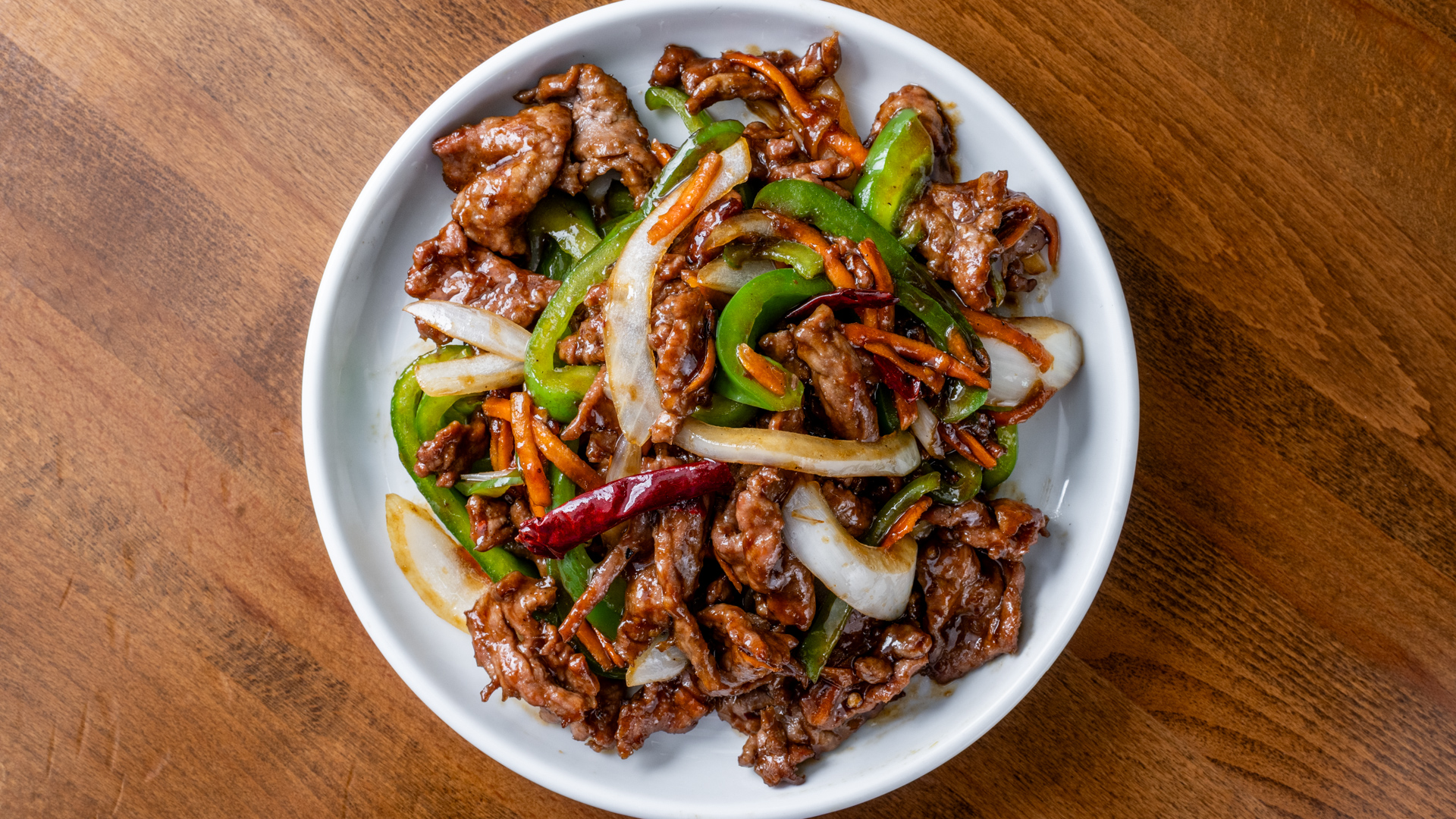 Top‑down view of glossy stir‑fried beef strips with green bell pepper, white onion, carrot strips and dried red chili in savory brown sauce, served in white bowl on wooden table.