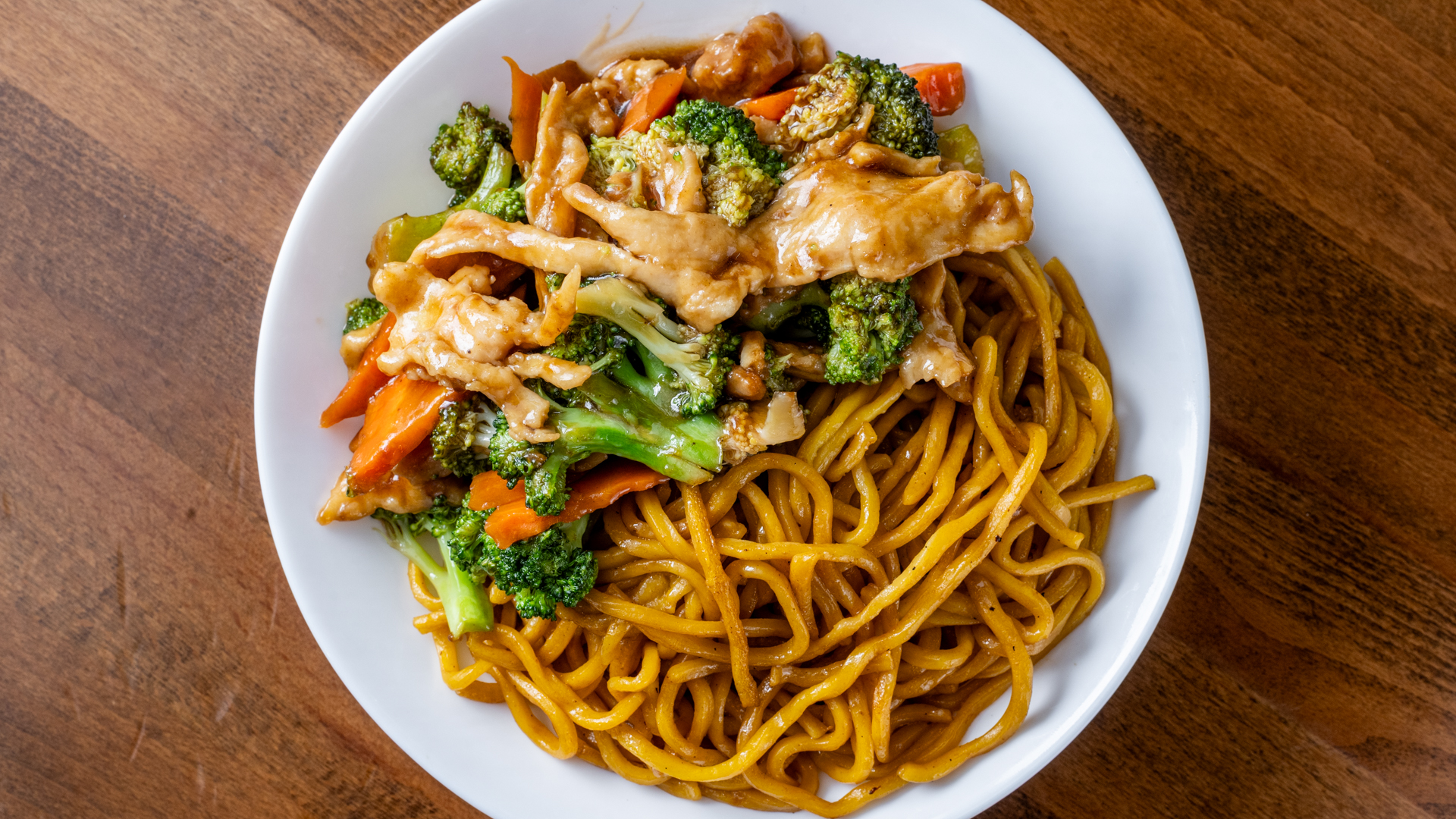 Overhead photo of chicken lo mein on white plate, thick egg noodles served with tender sauced chicken slices, broccoli florets and carrot pieces over dark wood table.