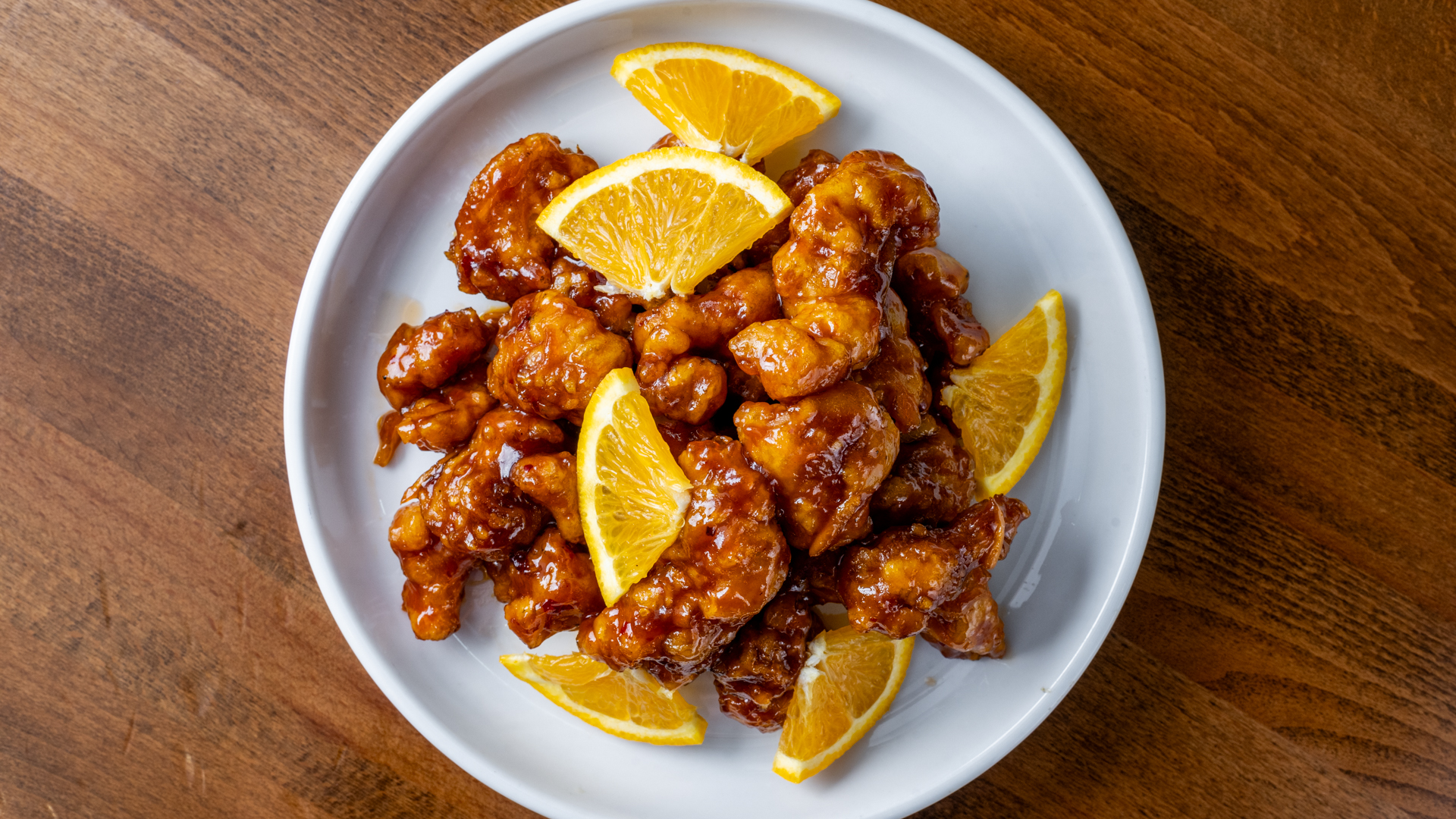 Top‑down shot of crispy fried orange chicken chunks coated in sticky sweet sauce, decorated with fresh orange wedges on round white plate against wooden background.