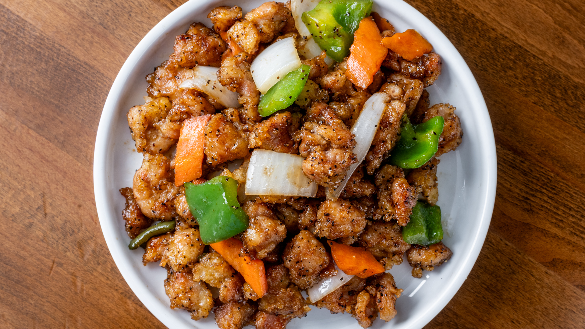 Overhead view of seasoned crispy black pepper diced chicken stir‑fry mixed with green bell pepper, white onion and carrot chunks in white bowl on dark wooden table.