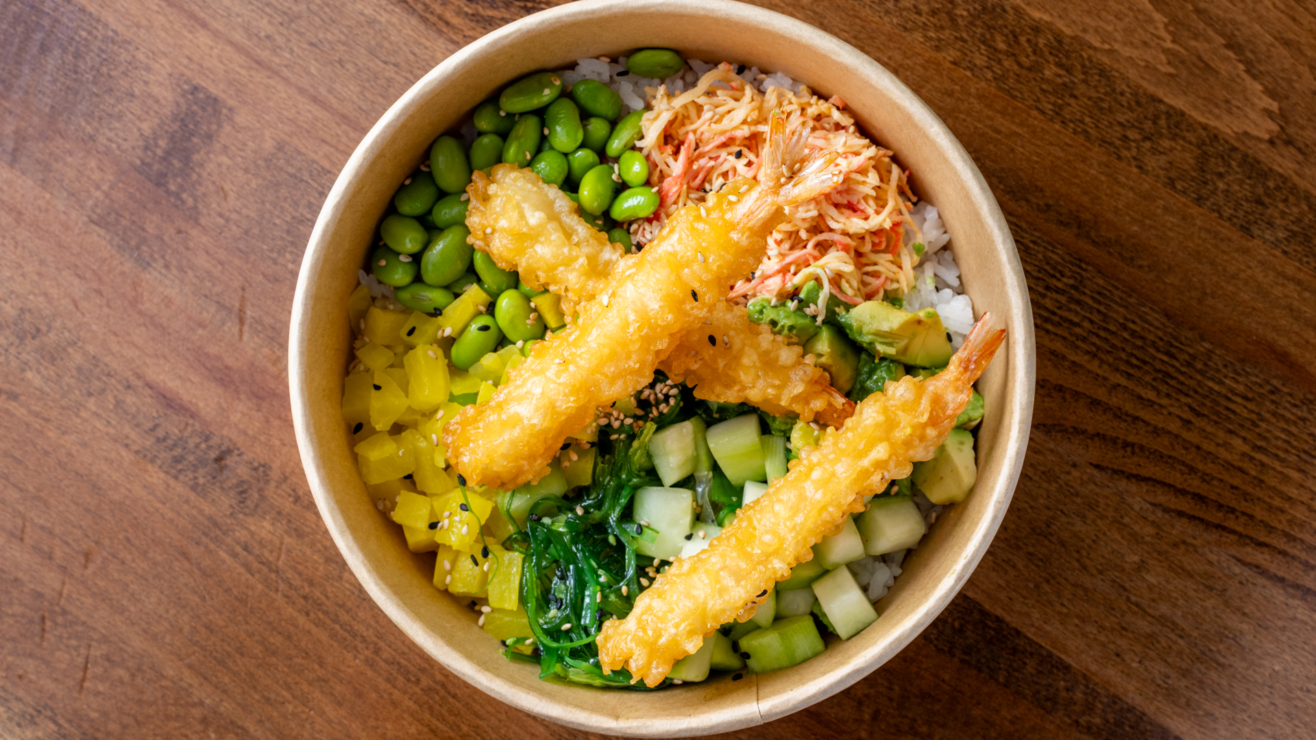 Top‑down tempura shrimp poke bowl in kraft paper bowl, three crispy golden fried shrimp, edamame, avocado, cucumber, seaweed salad, shredded crab and diced pepper over rice.