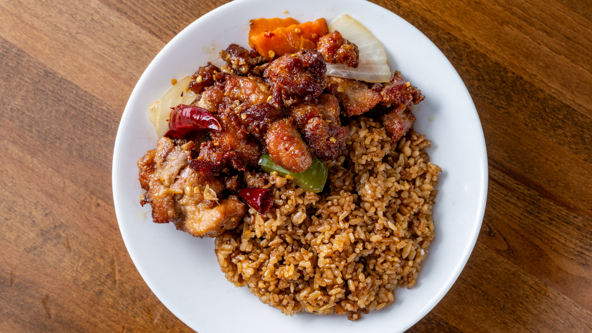 Over‑head view of spicy glazed fried chicken pieces with dried chili, onion and bell pepper, served alongside portion of brown fried rice on white plate on wooden table.