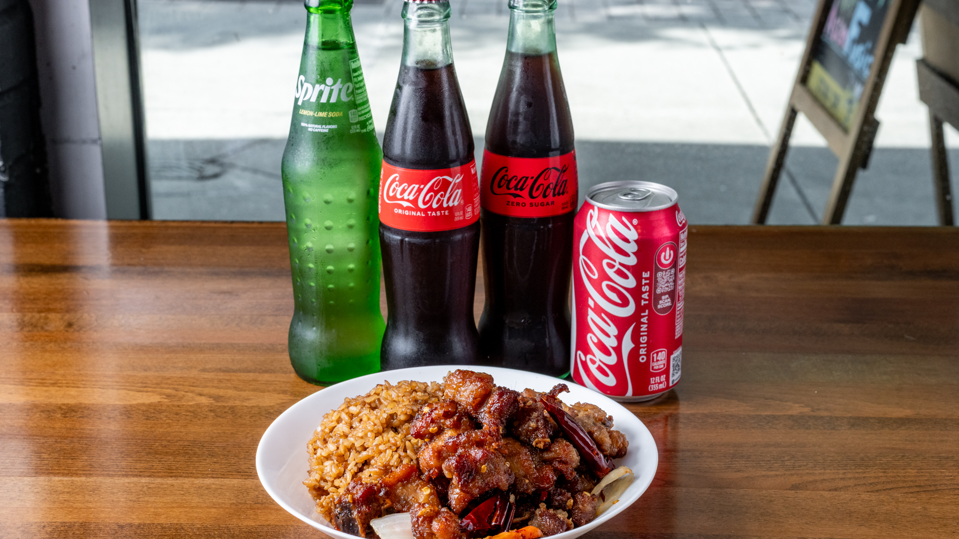 White bowl holding spicy glazed meat over fried rice, arranged with glass Sprite, Coca‑Cola, Coke Zero bottles and a Coke can on warm wooden table, outdoor window background.