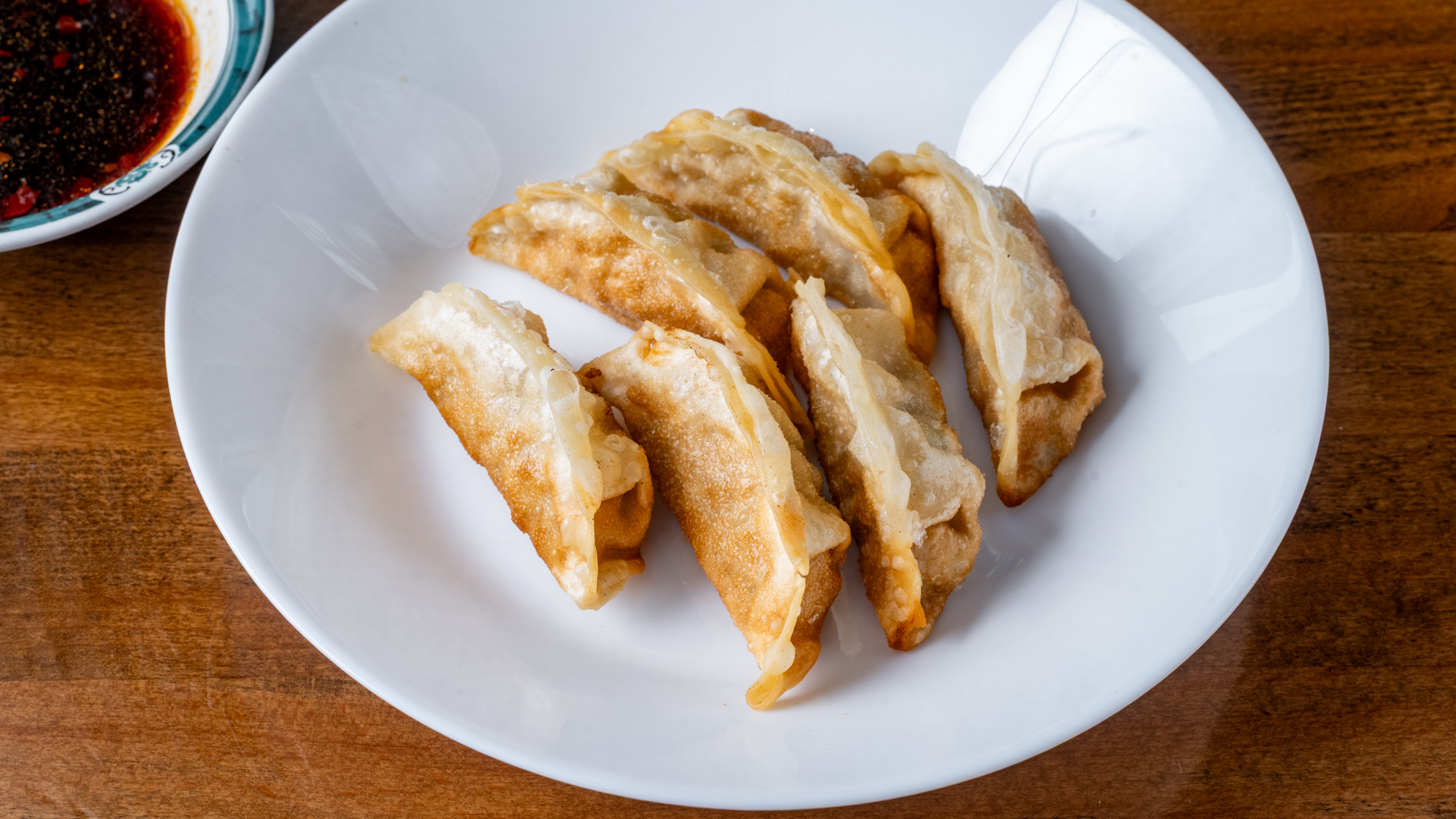Top‑down view of six crispy golden‑brown pan‑fried dumplings resting on white ceramic plate; spicy dipping sauce bowl sits at upper left corner on wooden table.