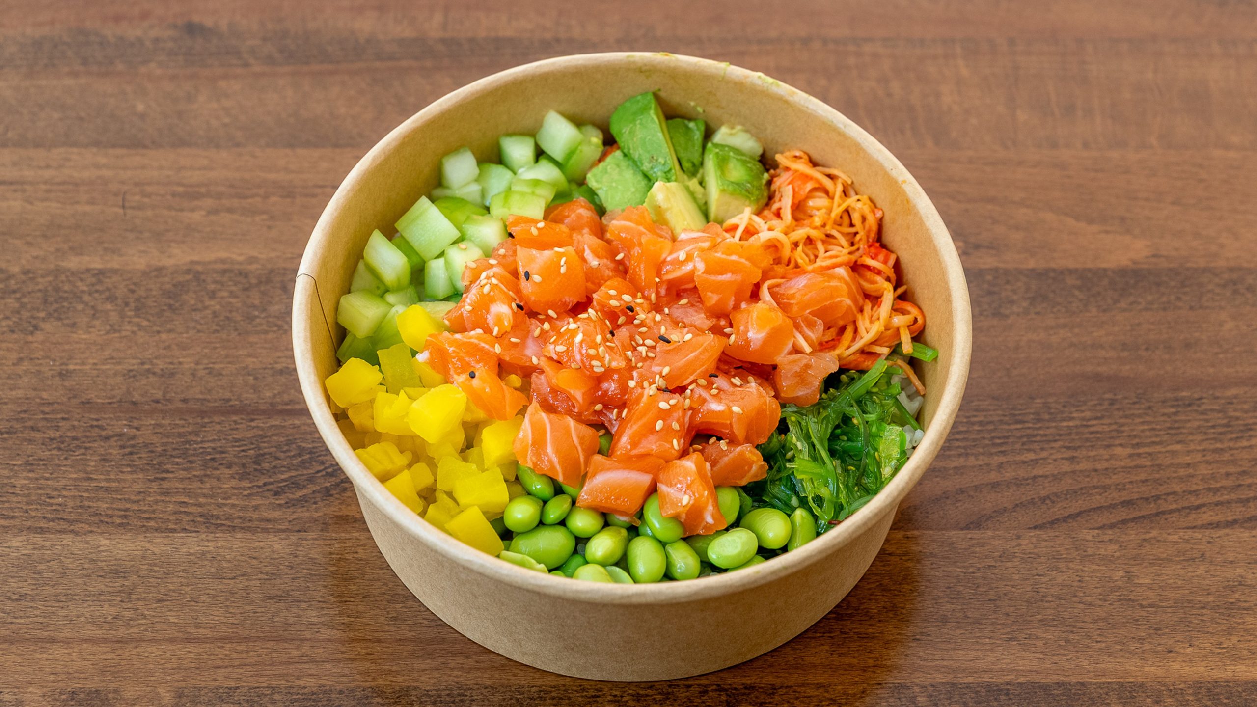 Paper poke bowl filled with raw salmon cubes, cucumber, mango, avocado, edamame, seaweed salad, shredded carrot, sprinkled white sesame seeds over rice base on wooden table.