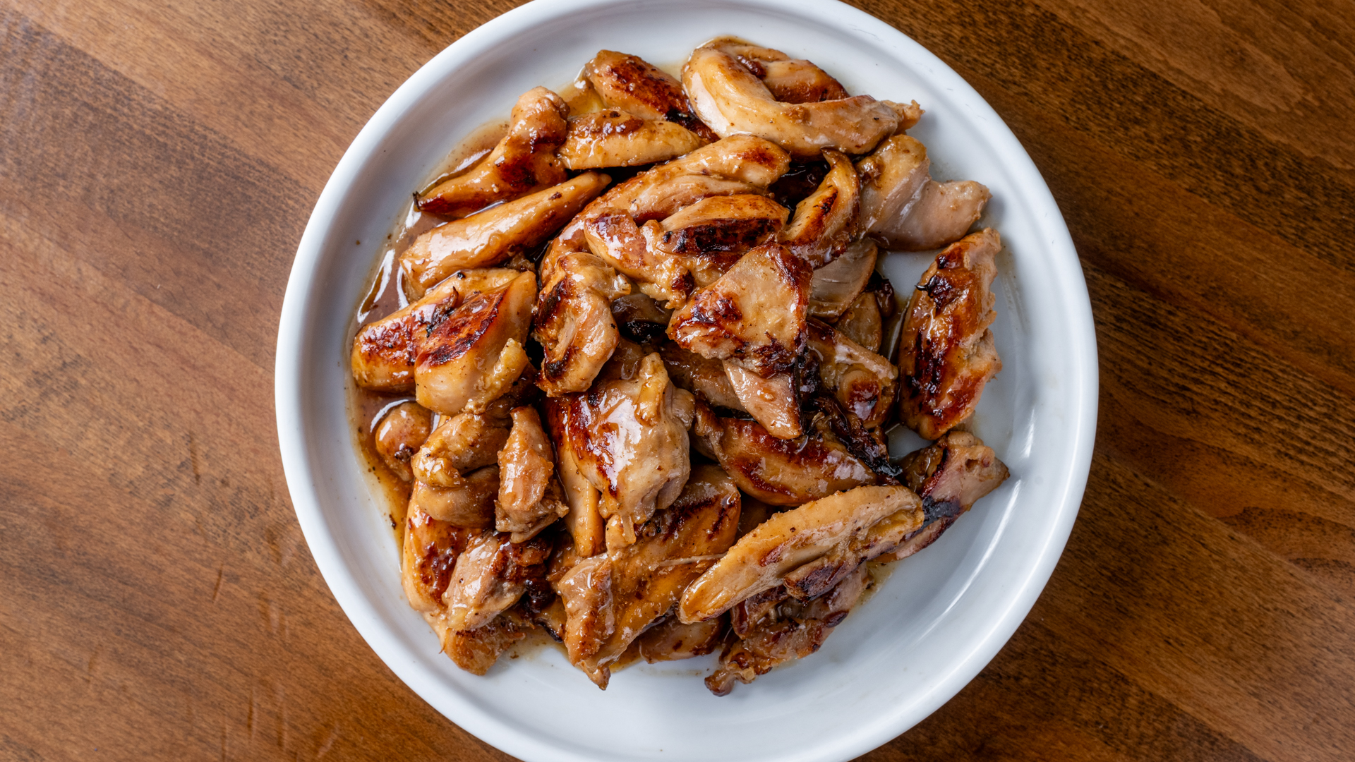 Overhead shot of tender grilled teriyaki chicken chunks with charred marks, glazed in rich brown sauce, piled inside round white ceramic plate set on dark wooden table.