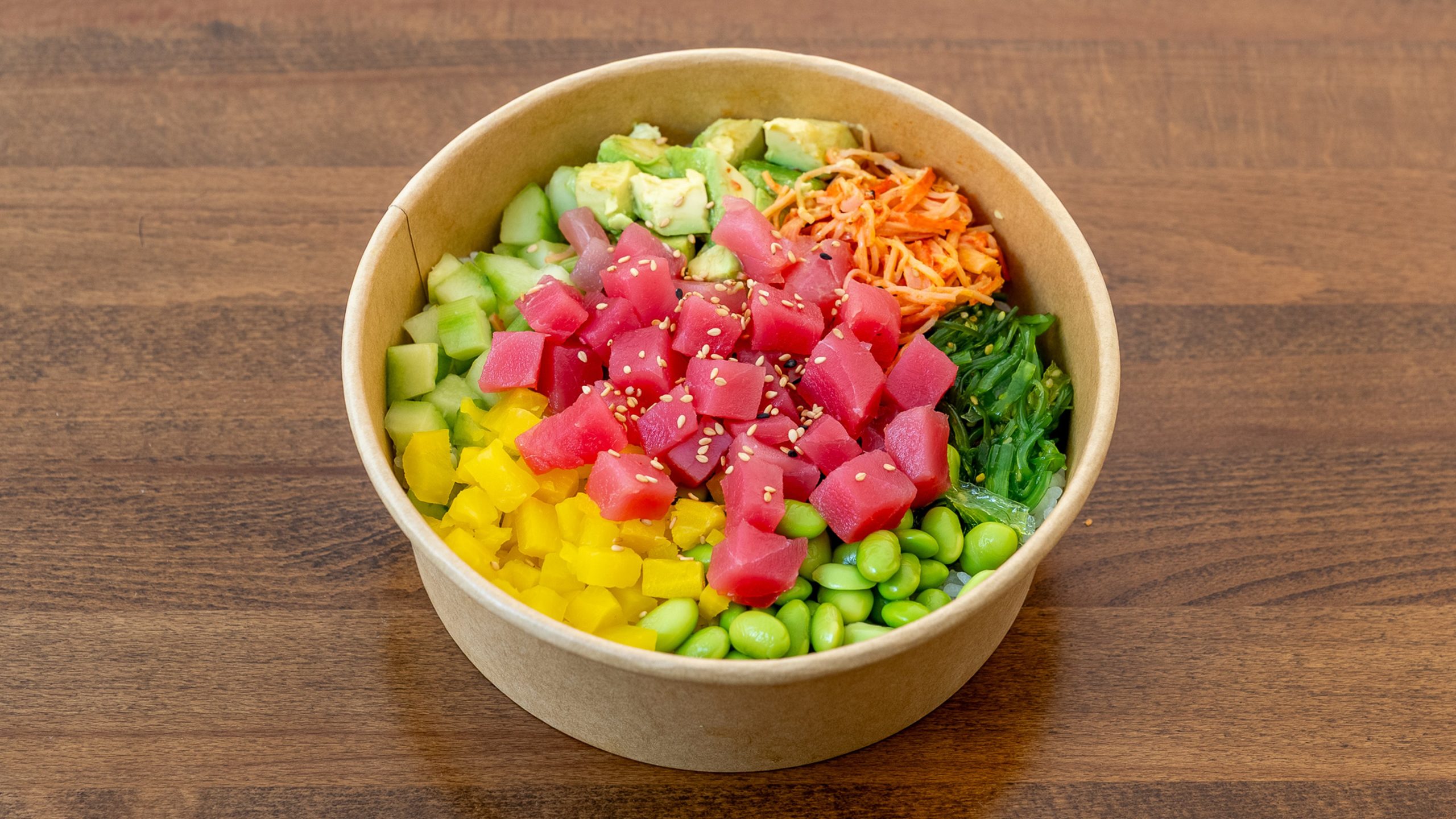 Kraft paper poke bowl with cubed raw tuna, avocado, cucumber, yellow bell pepper, edamame, seaweed salad, shredded carrot, sesame seeds layered over rice on wood table.