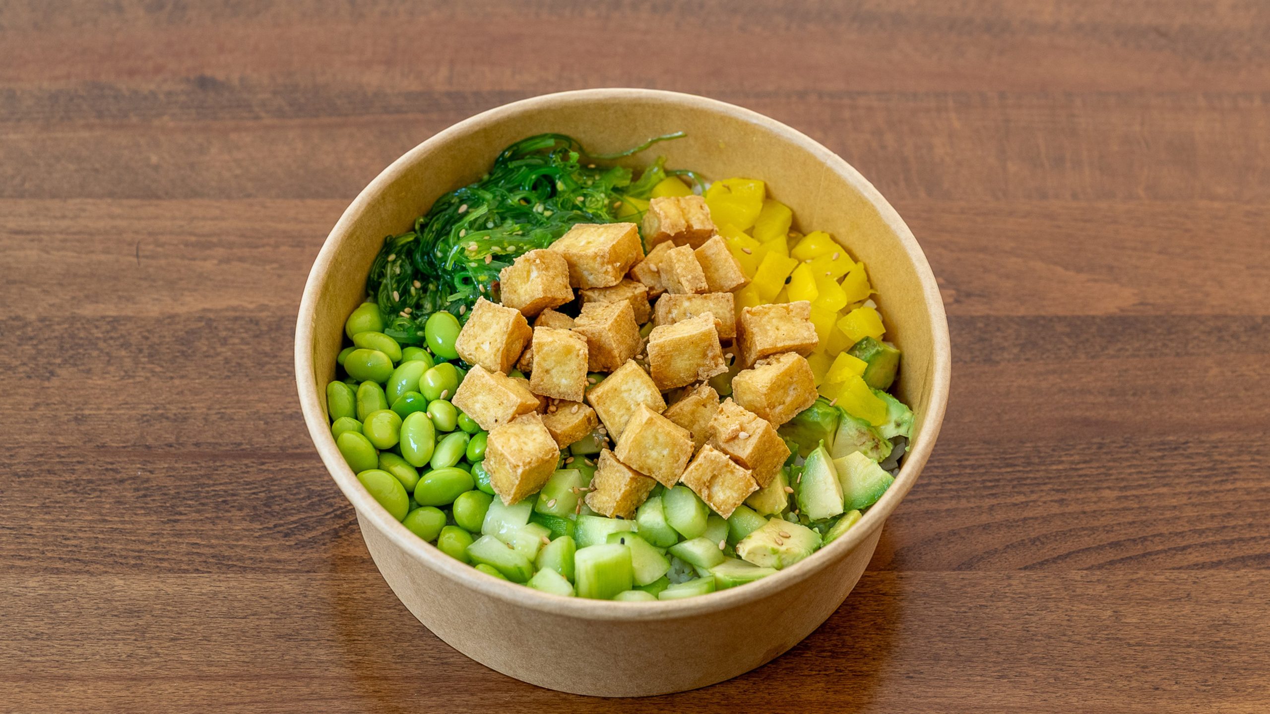 Paper poke bowl featuring golden fried tofu, edamame, cucumber, avocado, yellow bell pepper and seaweed salad, colorful plant‑based ingredients arranged over rice base on wooden table.