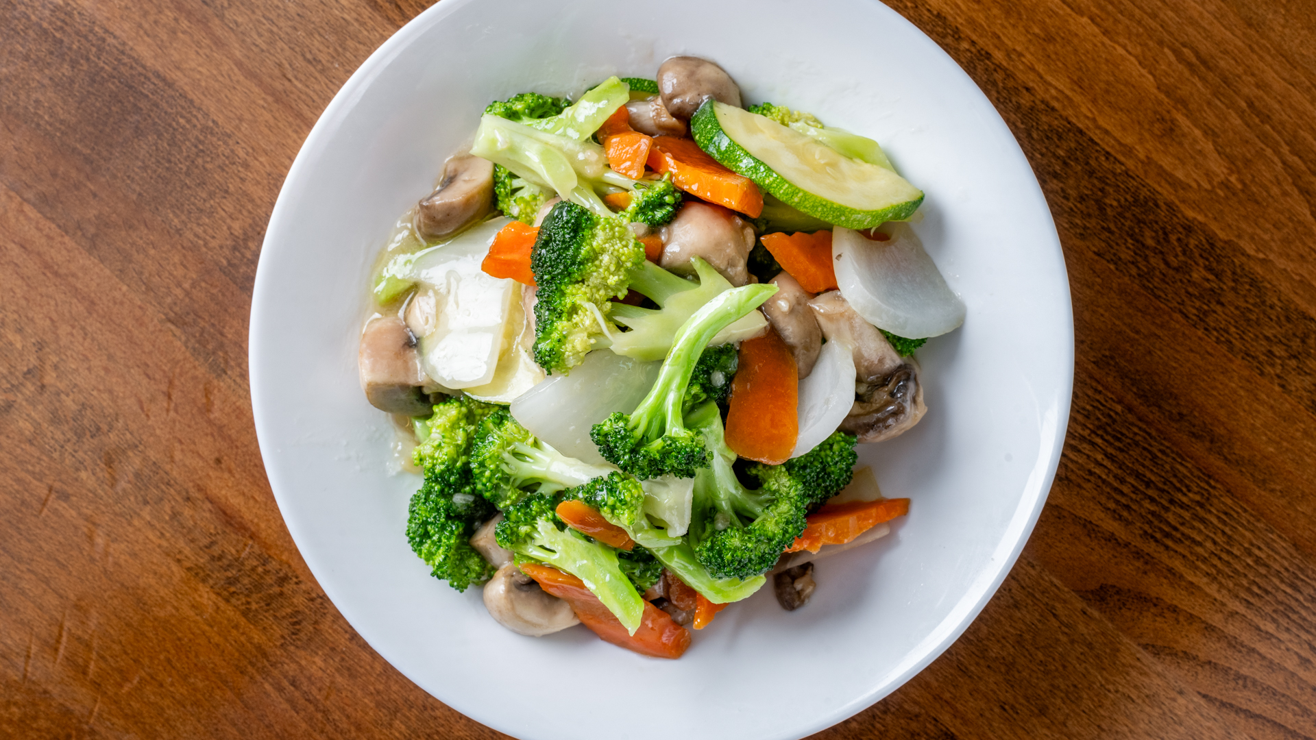 Top‑down photograph of stir‑fried vegetable medley including broccoli, mushroom, zucchini, carrot, onion coated in light sauce, served on white plate over wooden table surface.