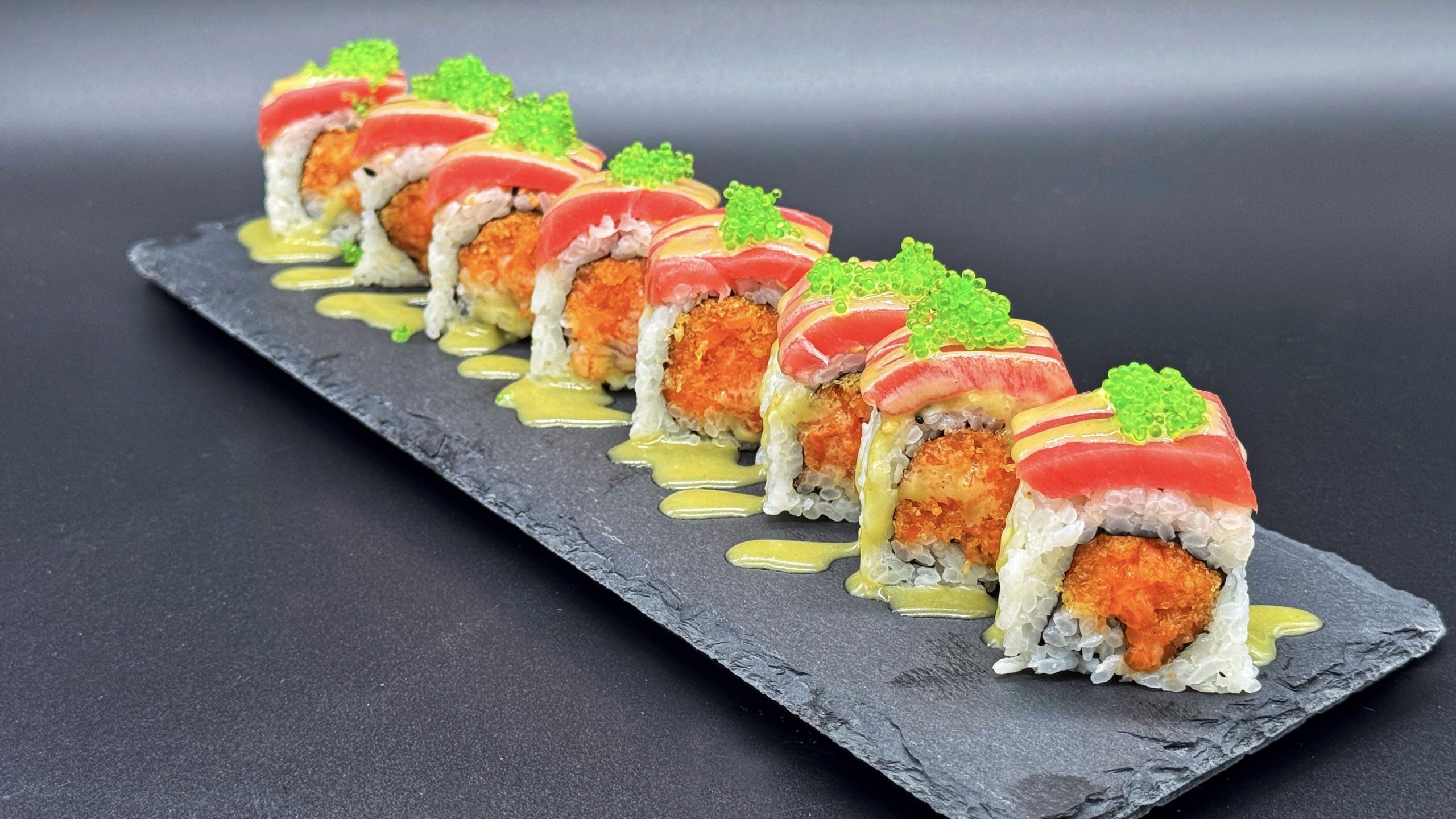Sliced sushi roll arranged on dark slate plate, topped with fresh tuna slices and bright green tobiko, creamy pale yellow sauce drizzled alongside against dark gray background.