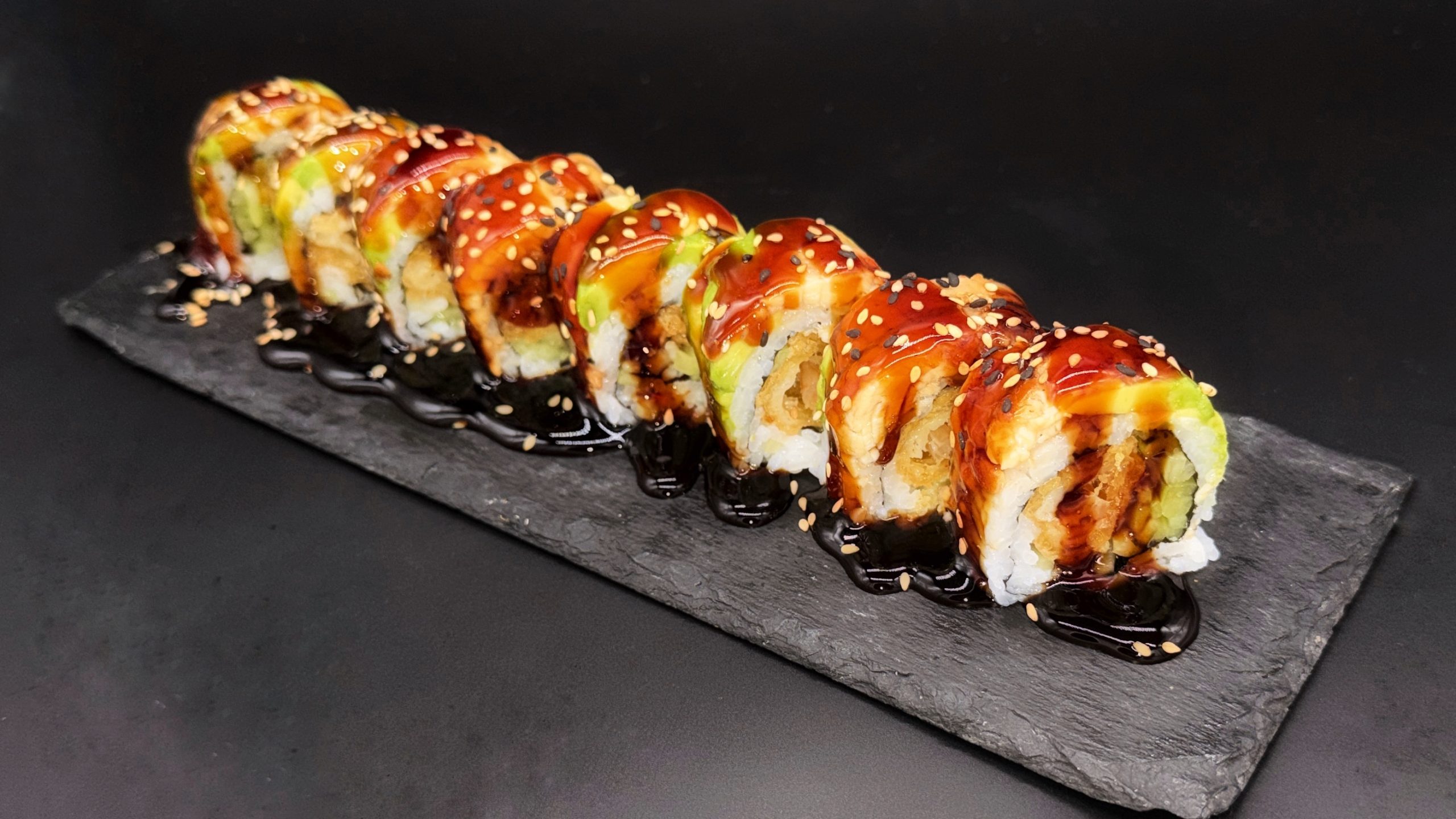 Portion of sushi roll slices laid on dark slate plate, glazed with thick dark eel sauce, sprinkled with black and white sesame seeds over avocado and tempura fillings.