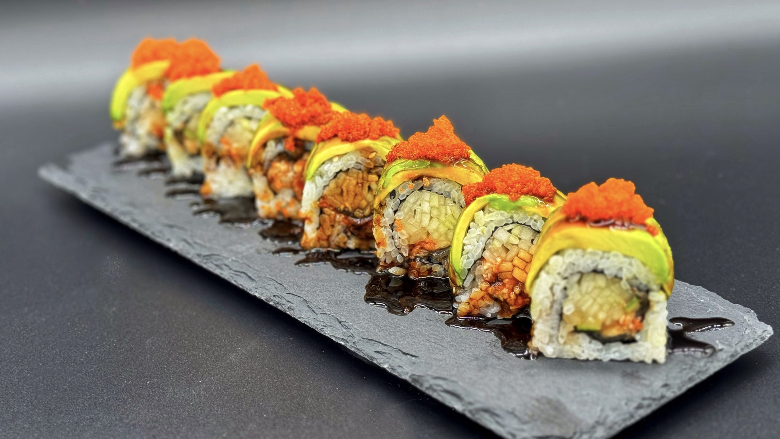 Row of sushi roll pieces on dark slate plate, topped with fresh avocado slices and bright orange tobiko, dark soy sauce drizzled across the plate surface.