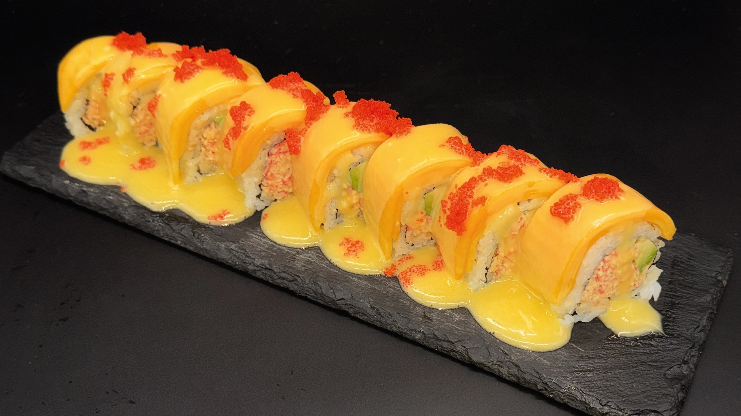 Sushi roll covered with fresh mango slices, creamy yellow sauce poured over, garnished with red tobiko roe, served on dark slate plate against black background.